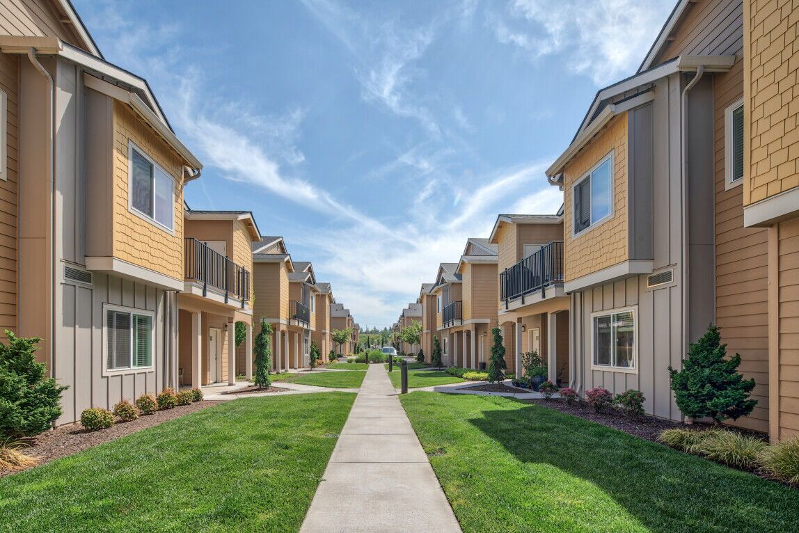 Rows of townhouses with beige siding and green lawns flank a concrete pathway under a blue sky.