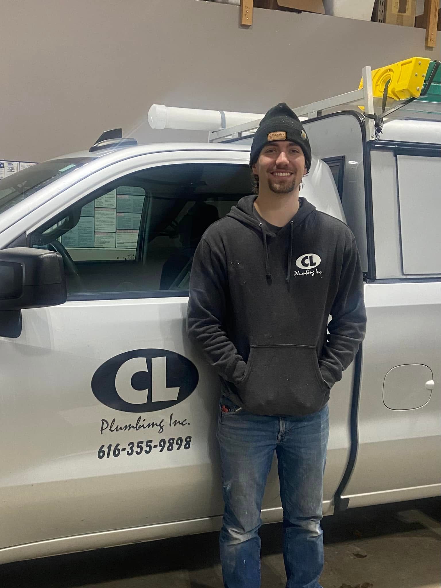 Man standing beside a silver company truck with a logo, wearing a black beanie and hoodie in a warehouse setting