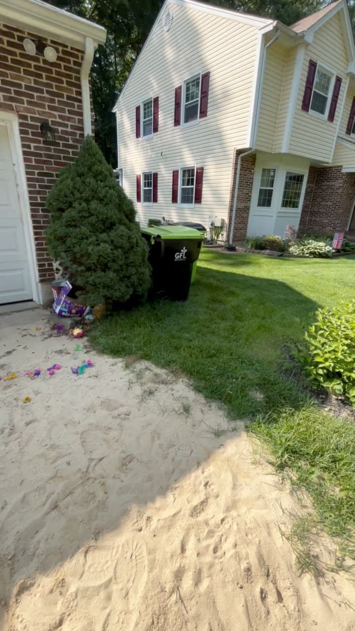 Backyard with sand, trash can, and house; green grass, beige house, brick garage.