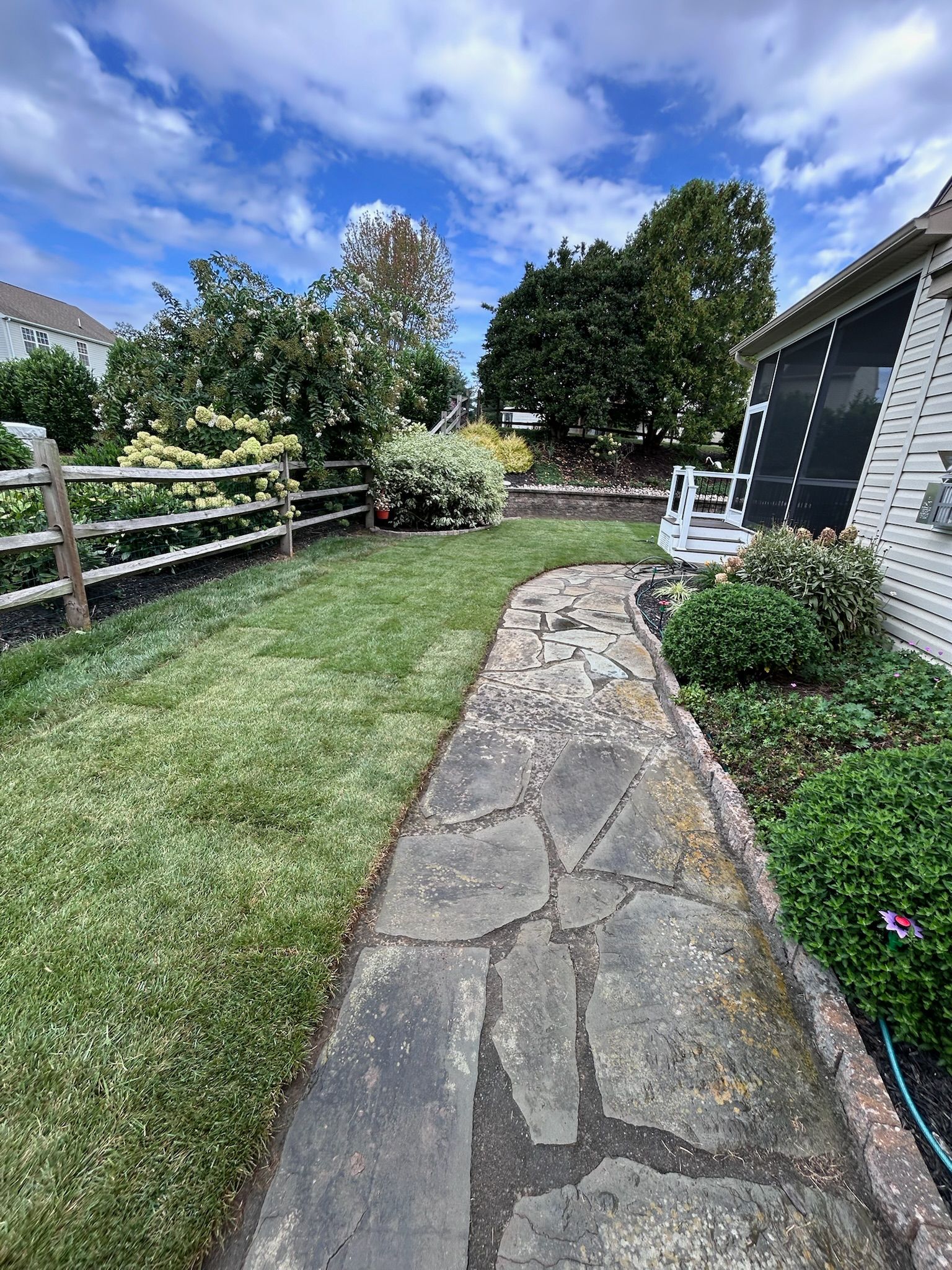 A stone walkway leads through a backyard toward trees and a split-rail fence under a blue, cloudy sky.