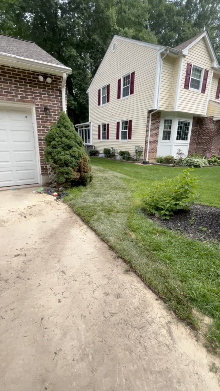 A driveway leads to a two-story beige house and a brick garage. Green lawn with plants runs alongside.