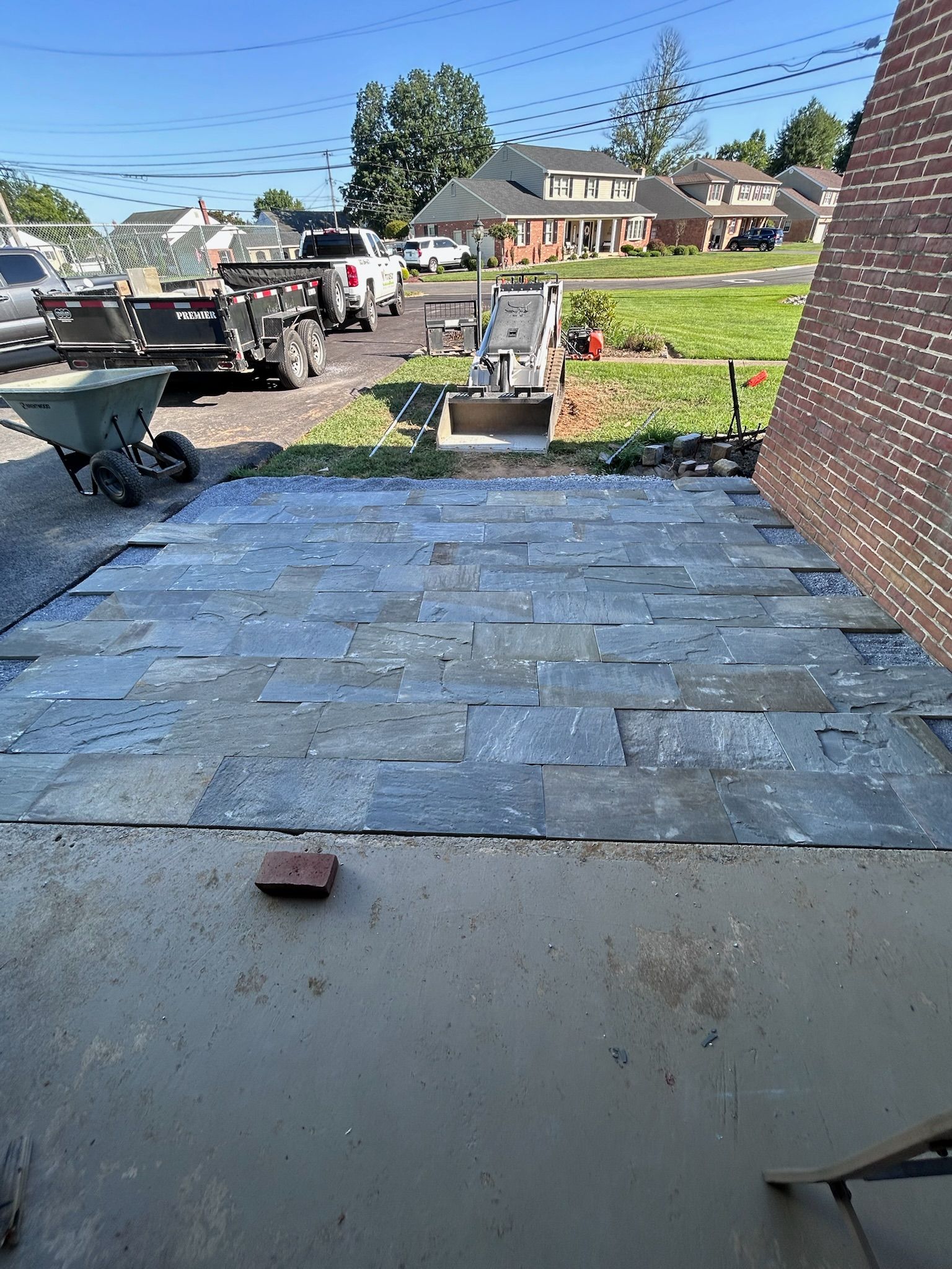 A new grey stone patio installation next to a brick building, with a wheelbarrow and construction equipment in the yard.