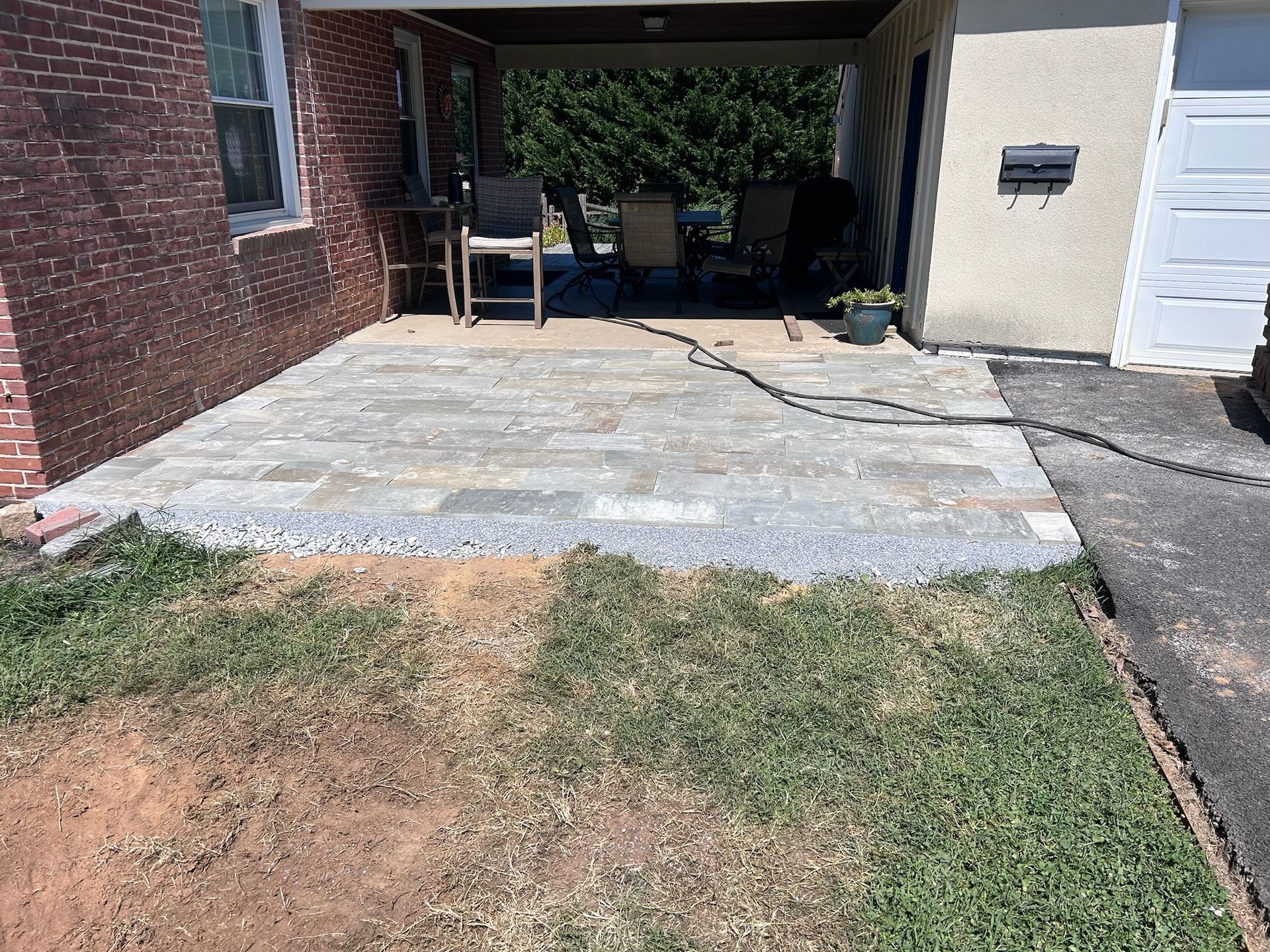 A paved stone patio with outdoor furniture under a covered porch next to a brick house and a paved driveway.