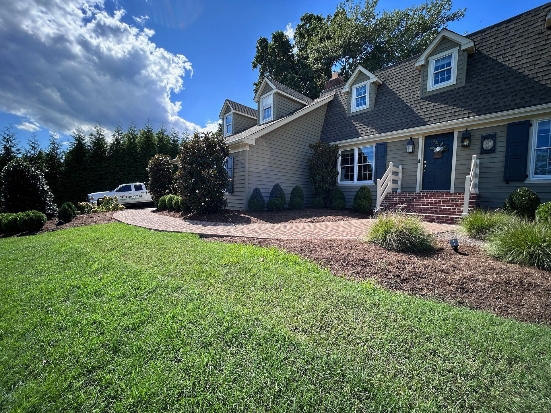 A tan house with a brick walkway and front dormers sits behind a lush green lawn under a bright blue sky.