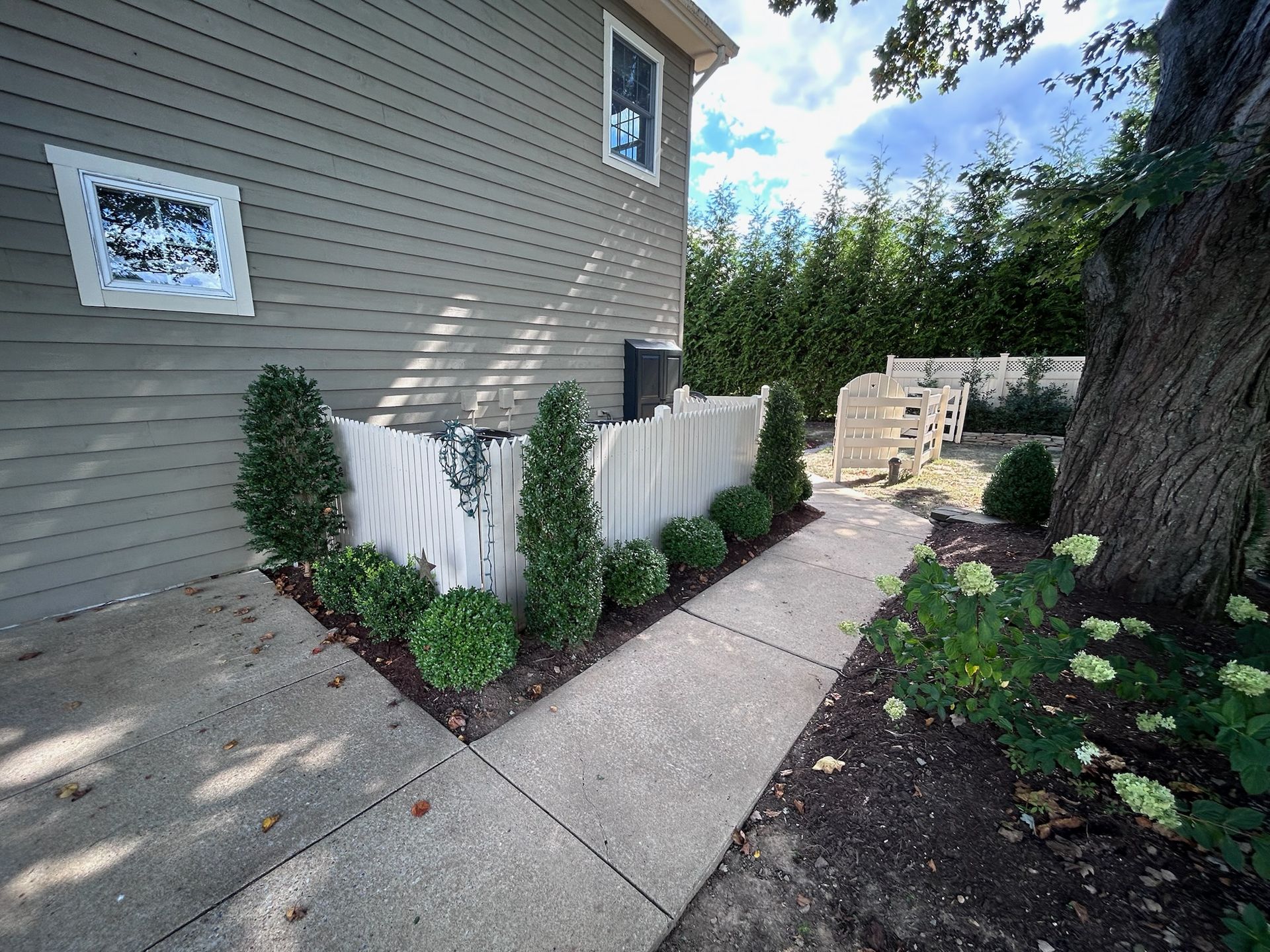 A white picket fence encloses a mechanical unit next to a house with beige siding, lined with small, manicured shrubs.