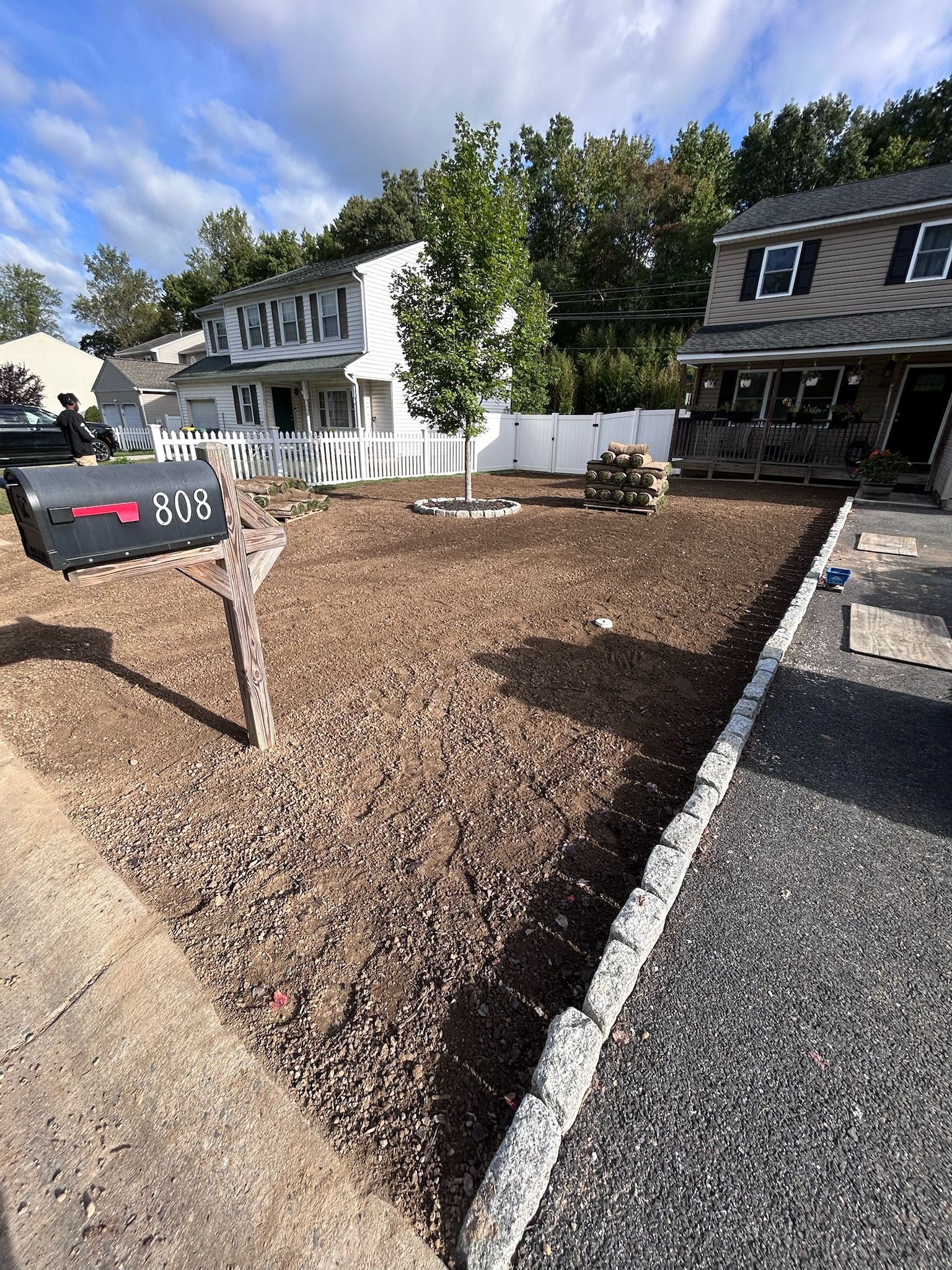 A view of a residential front yard covered in wood mulch, featuring a mailbox, a small tree, and a stone-edged driveway.
