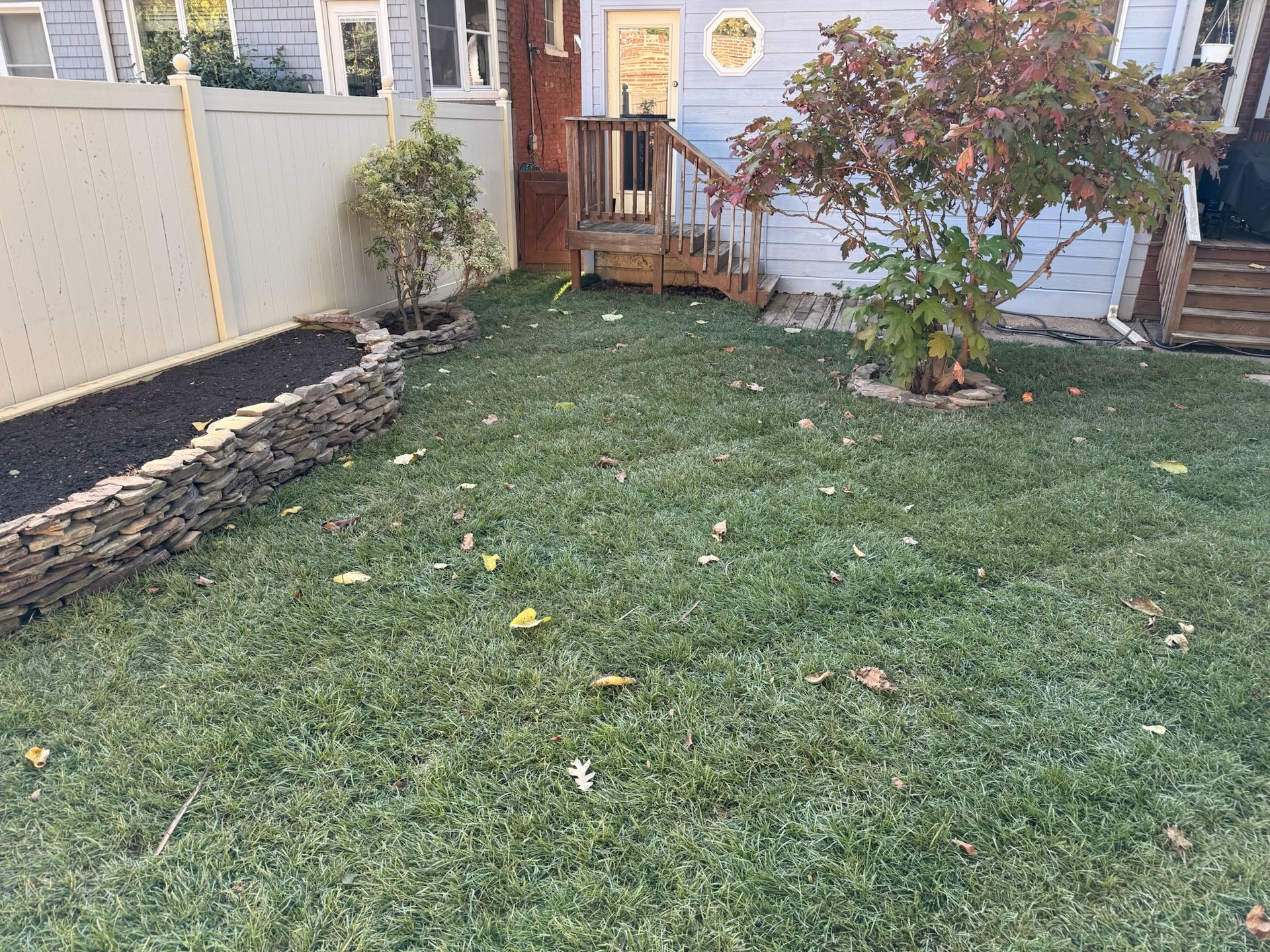A backyard with a stone-edged planter filled with dark mulch, a green lawn, and a small tree near house steps.