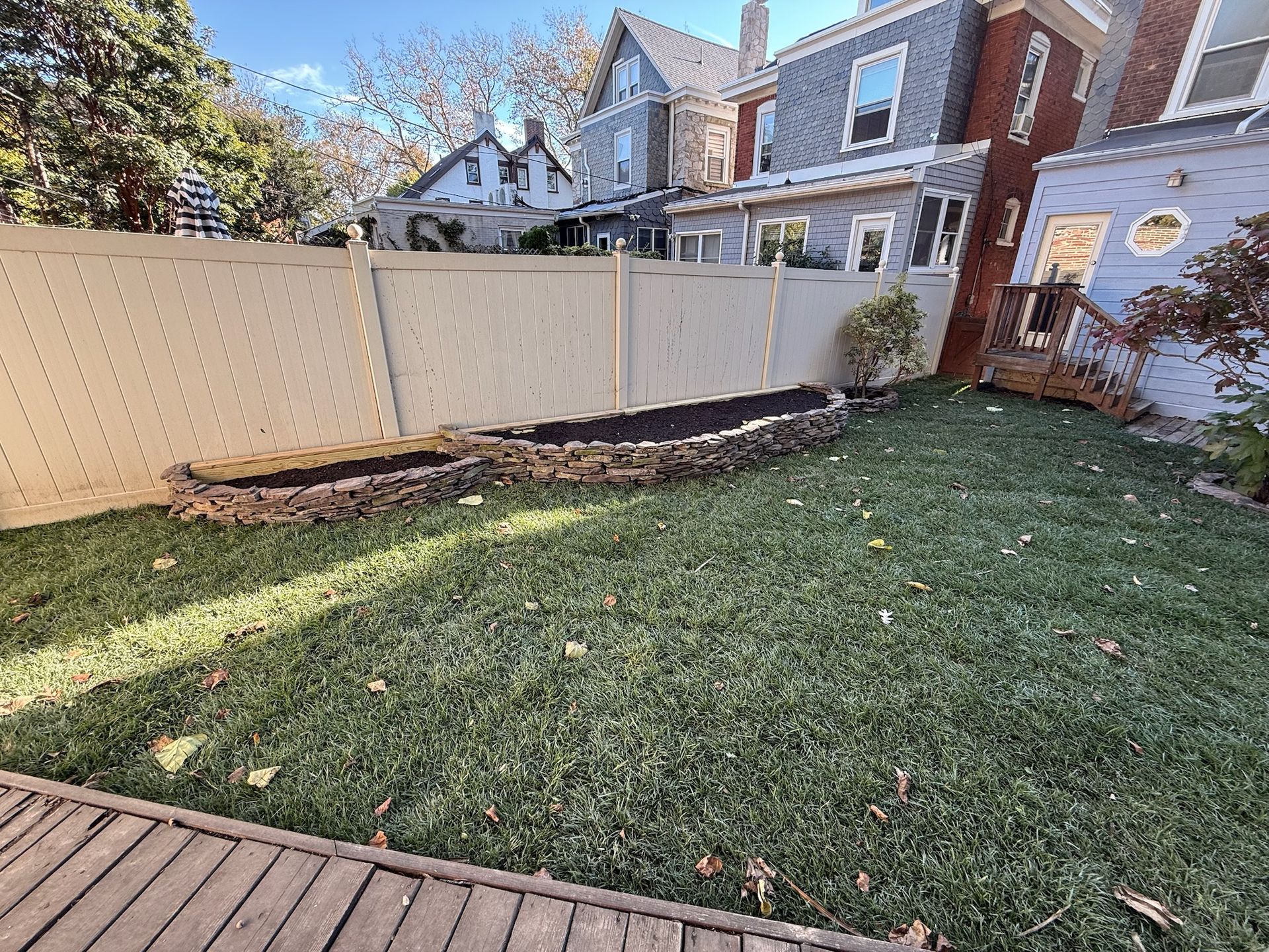 A fenced backyard lawn featuring a curved, stone-lined garden bed filled with dark soil, viewed from a wooden deck.