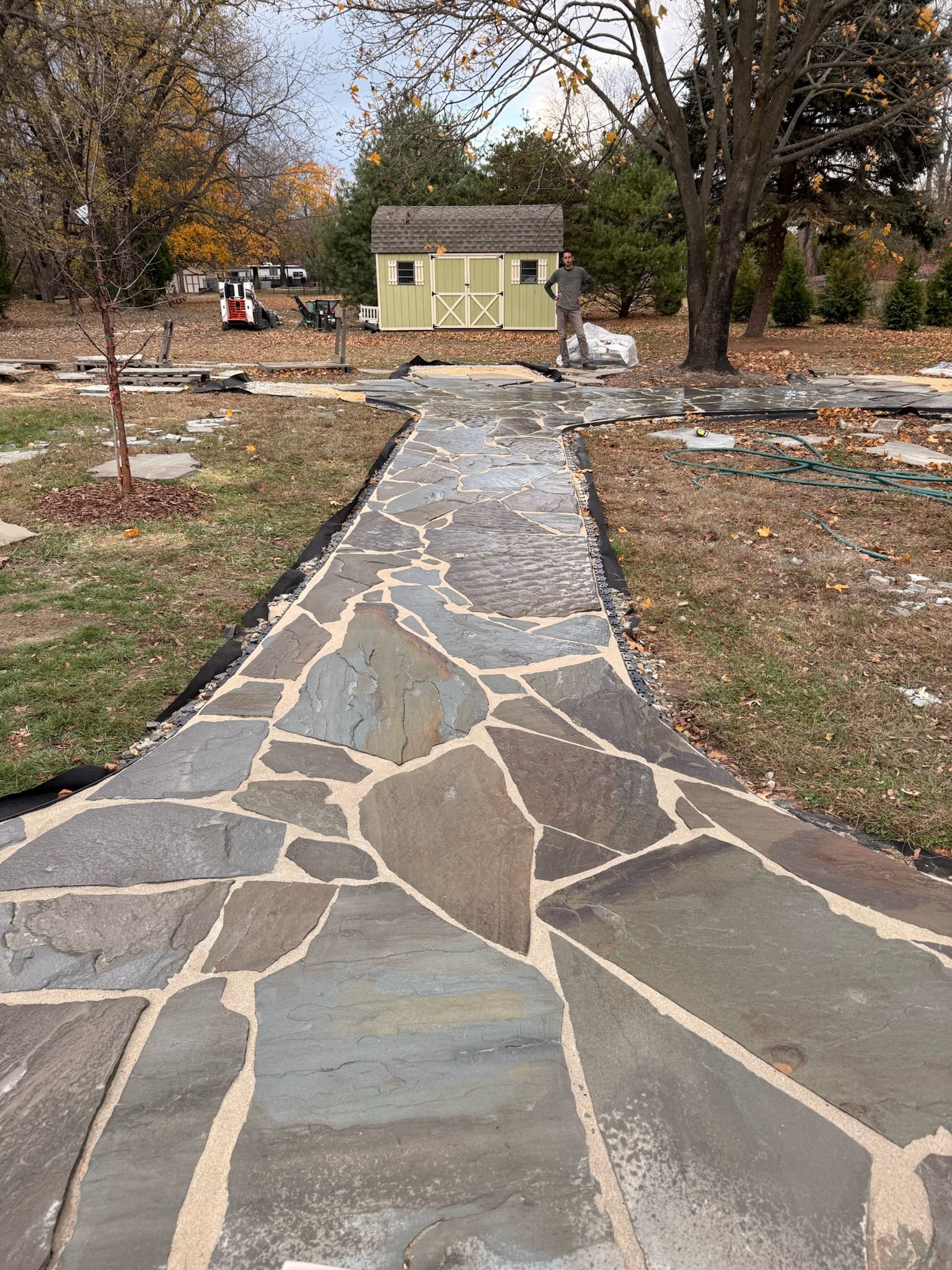 A stone flagstone walkway with light-colored grout leads through a grassy yard toward a small, light-green storage shed.