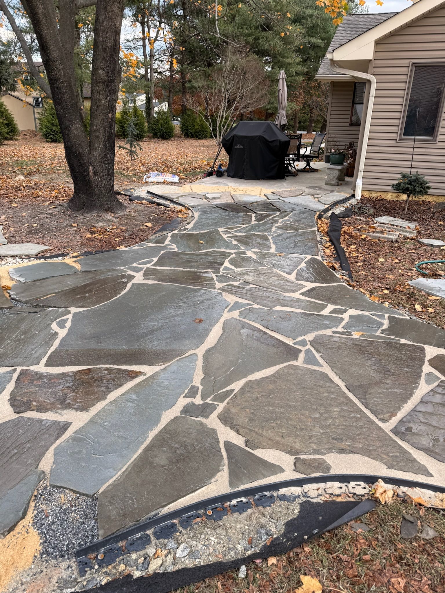 A flagstone patio walkway leads to a house exterior featuring a grill, surrounded by trees and fallen autumn leaves.