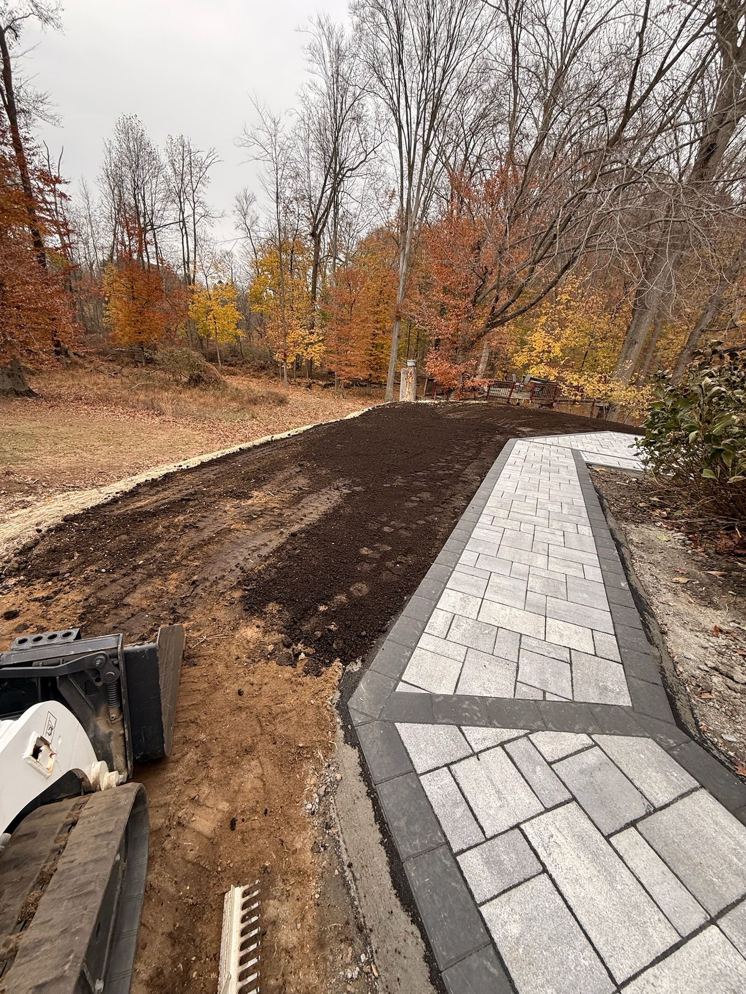 A gray paved walkway with a dark border runs through a yard, next to a strip of dark soil and a piece of construction equipment.