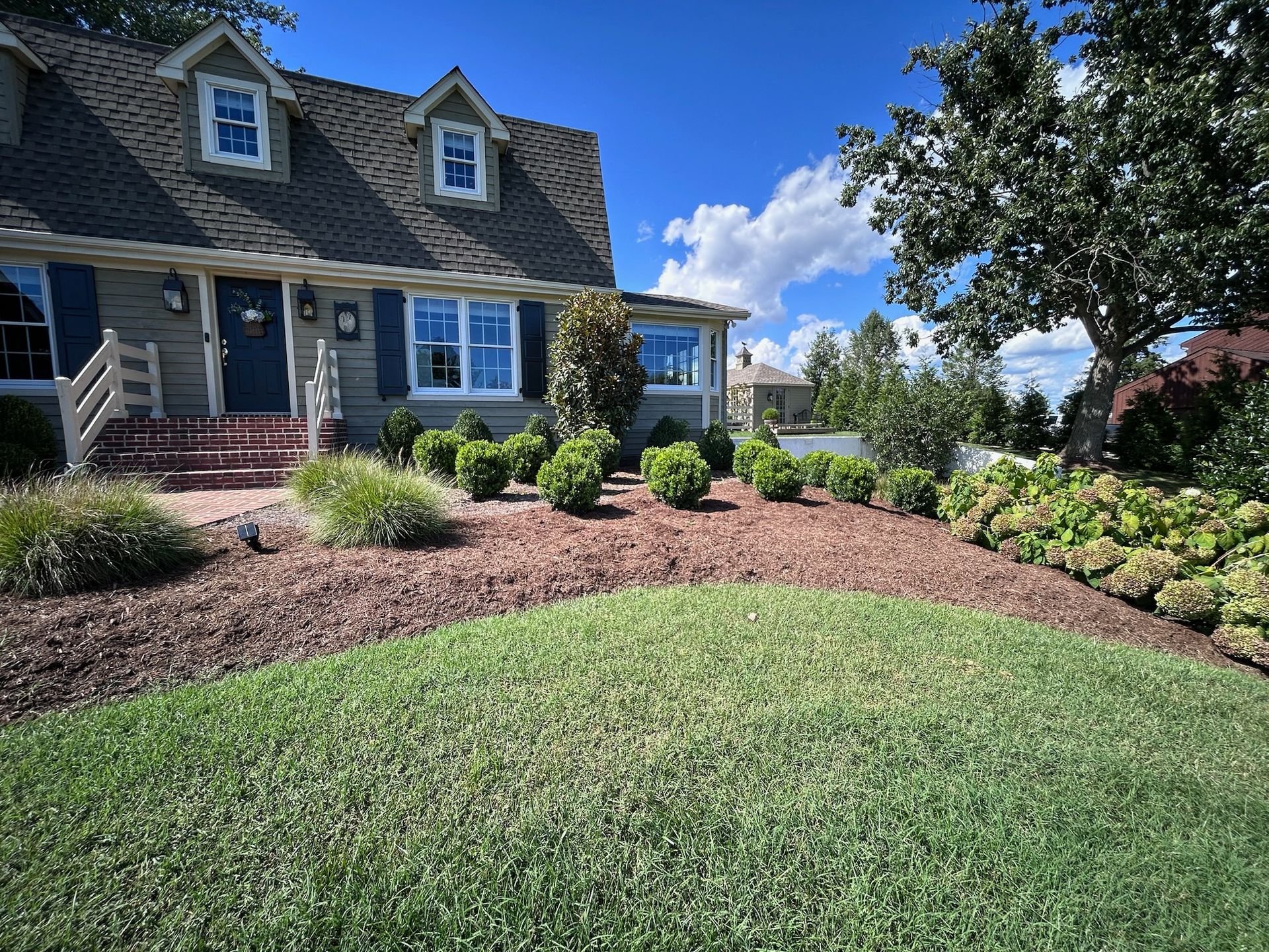 A grey house with dormer windows, blue shutters, and a front yard featuring a manicured lawn and mulched garden beds.