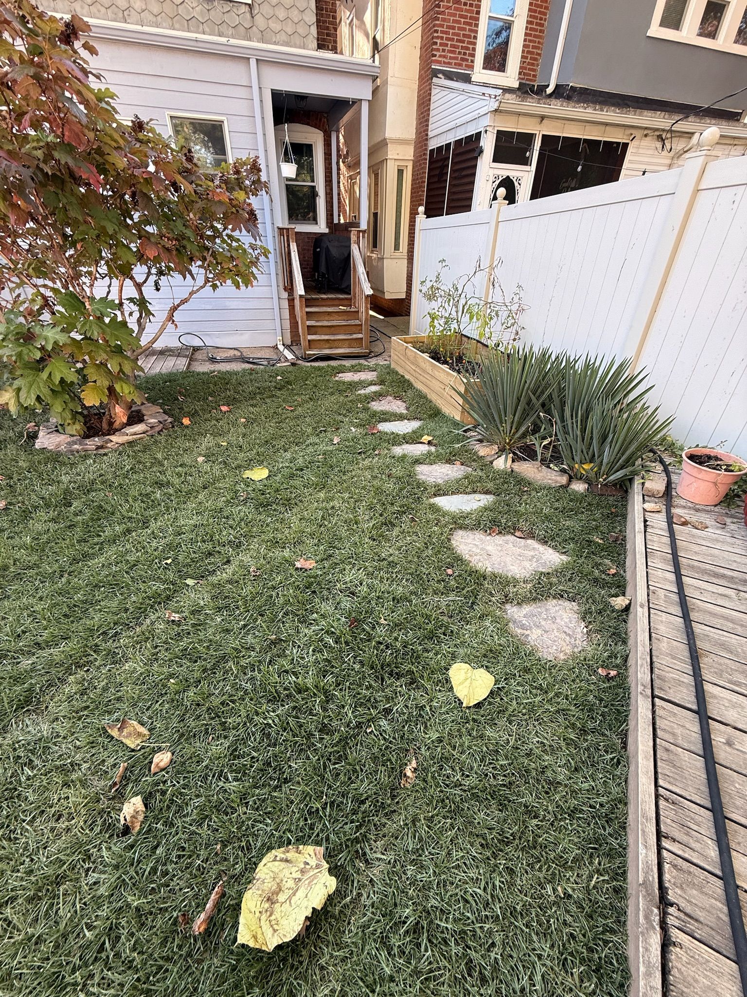 A backyard view featuring a lawn with a stone path leading toward wooden steps, a white fence, and a small house exterior.