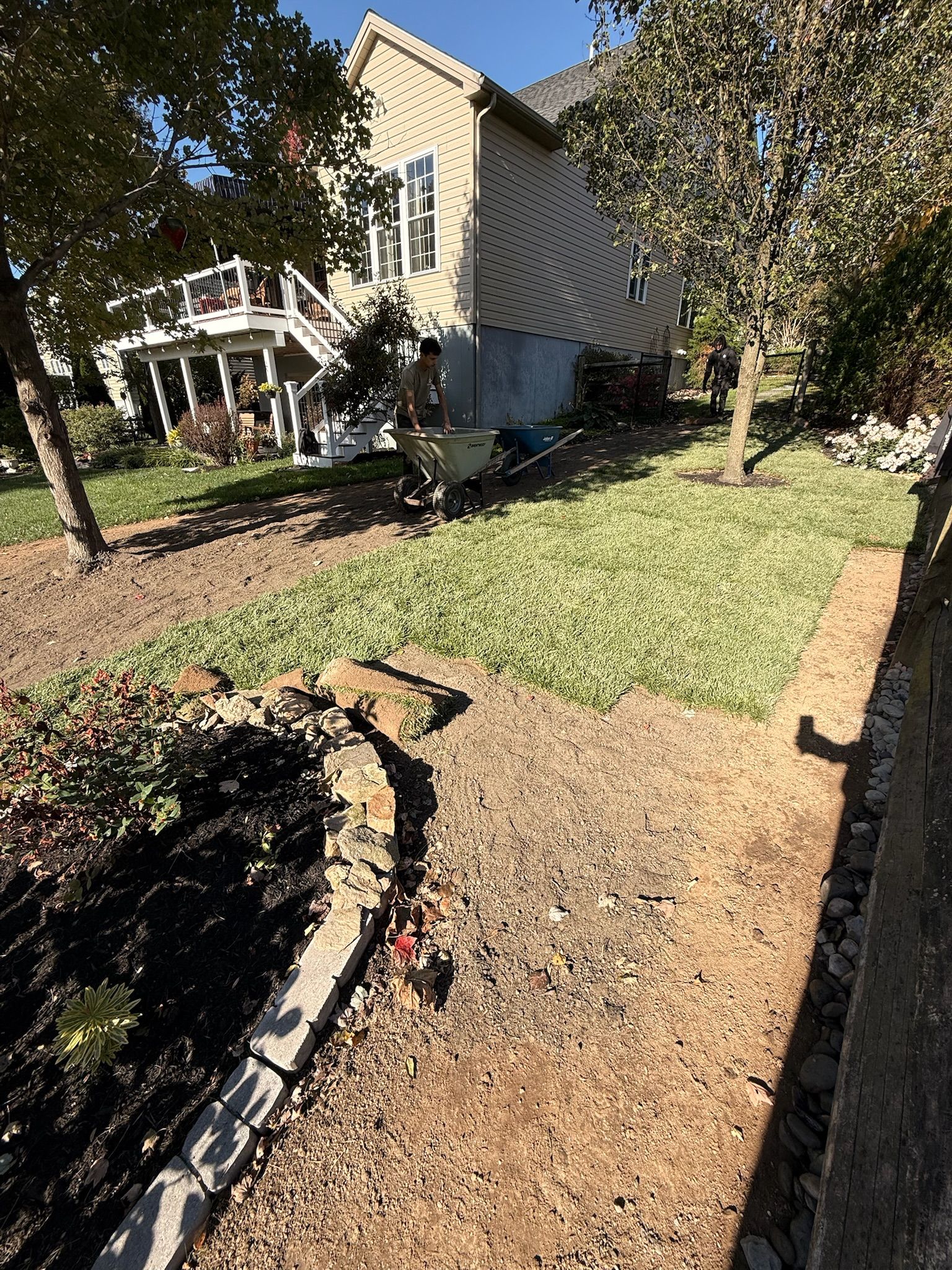 A person pushing a wheelbarrow through a sunlit yard with a house, landscaping, and tree-covered grass.