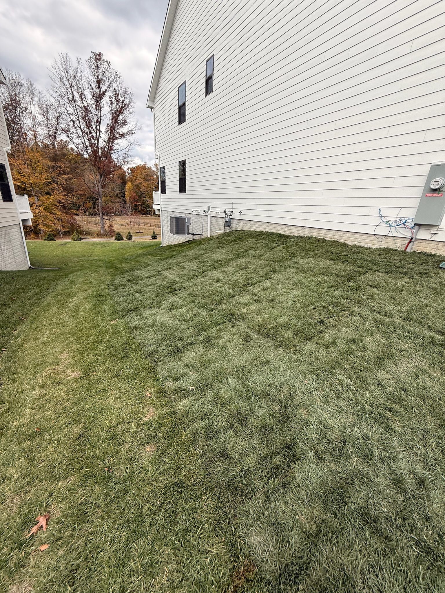 The side of a white house with horizontal siding overlooks a sloping green lawn and trees in the distance.