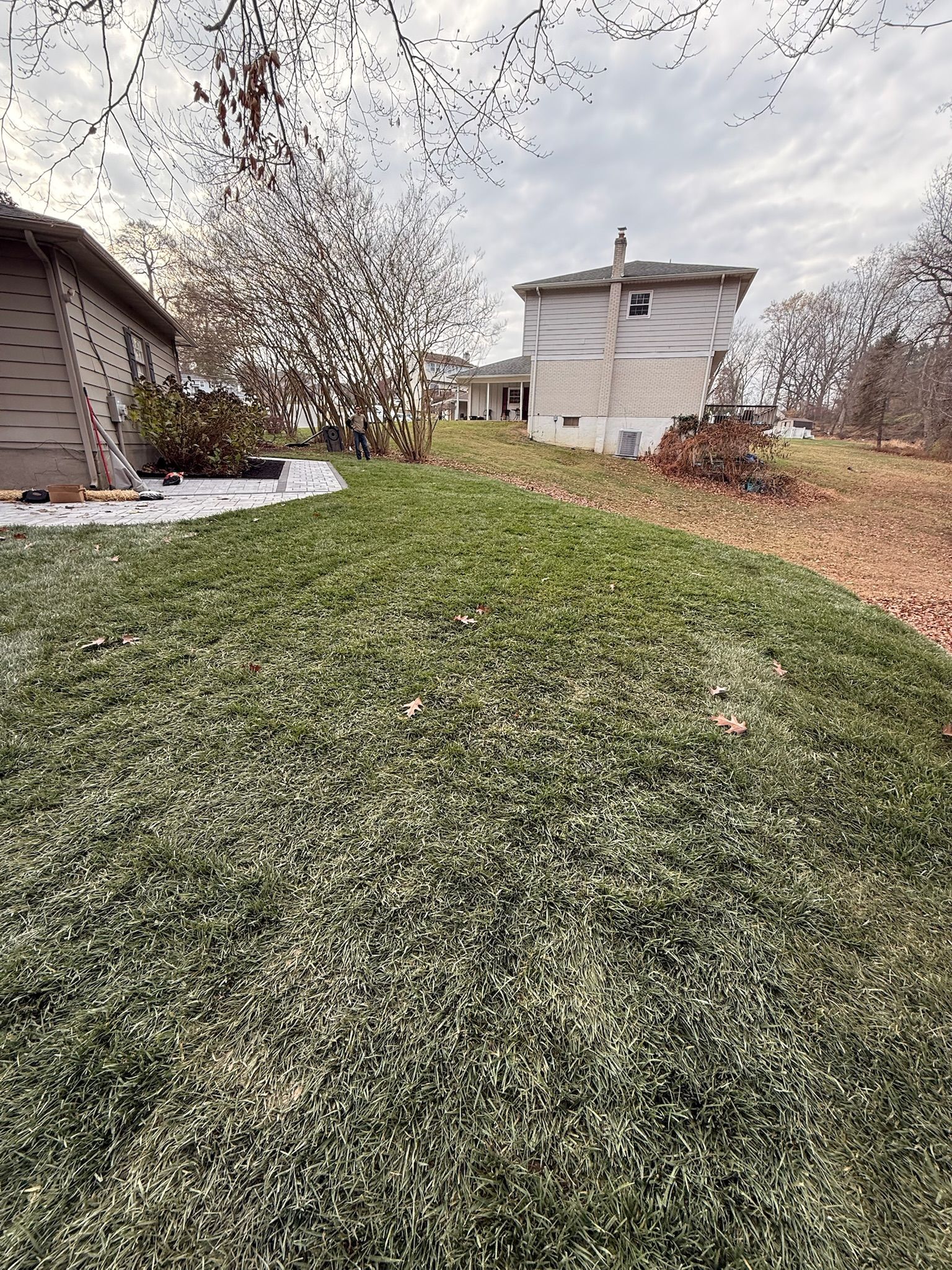 A view of a grassy yard sloping upward toward a white multi-story house under a cloudy sky.