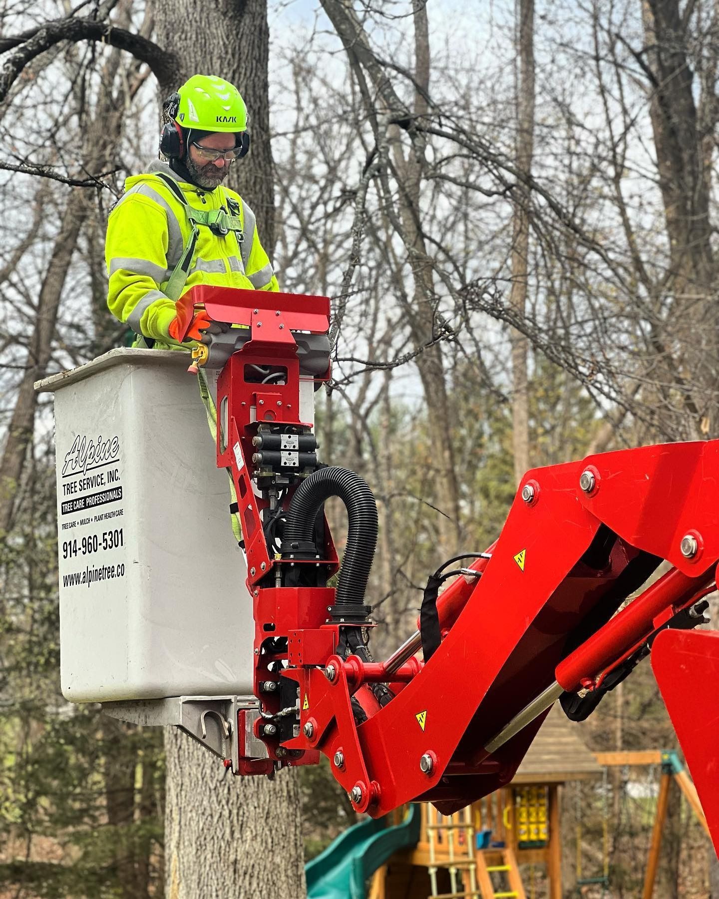 A person in safety gear and a bright yellow jacket operates a red aerial lift bucket in a wooded area.