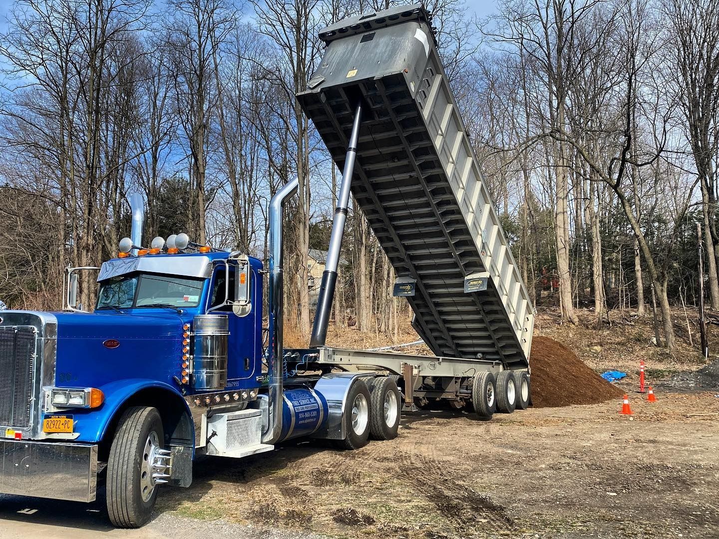 A blue semi-truck with its dump trailer raised, unloading a pile of soil on a dirt lot near a line of trees.