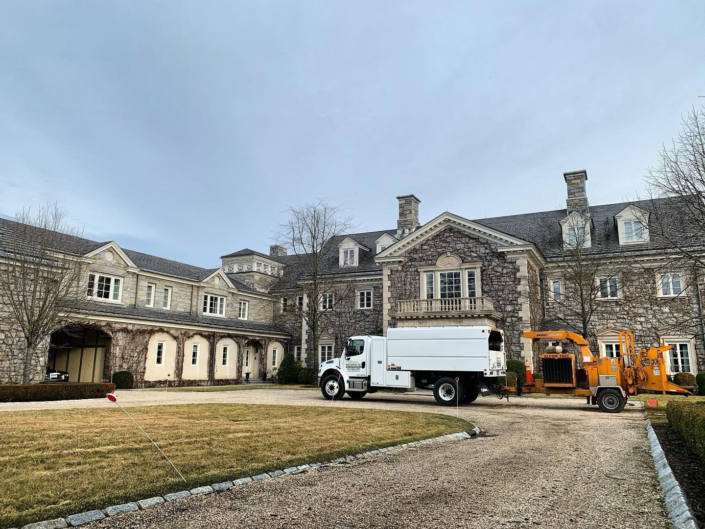 A stone mansion exterior with a parked white utility truck and an orange wood chipper on a gravel driveway.