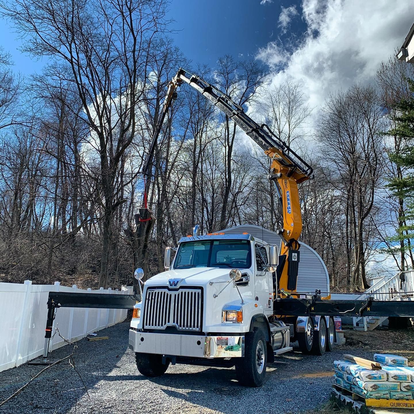 A white Western Star truck with a yellow crane boom extended, working on tree maintenance near a white vinyl fence.
