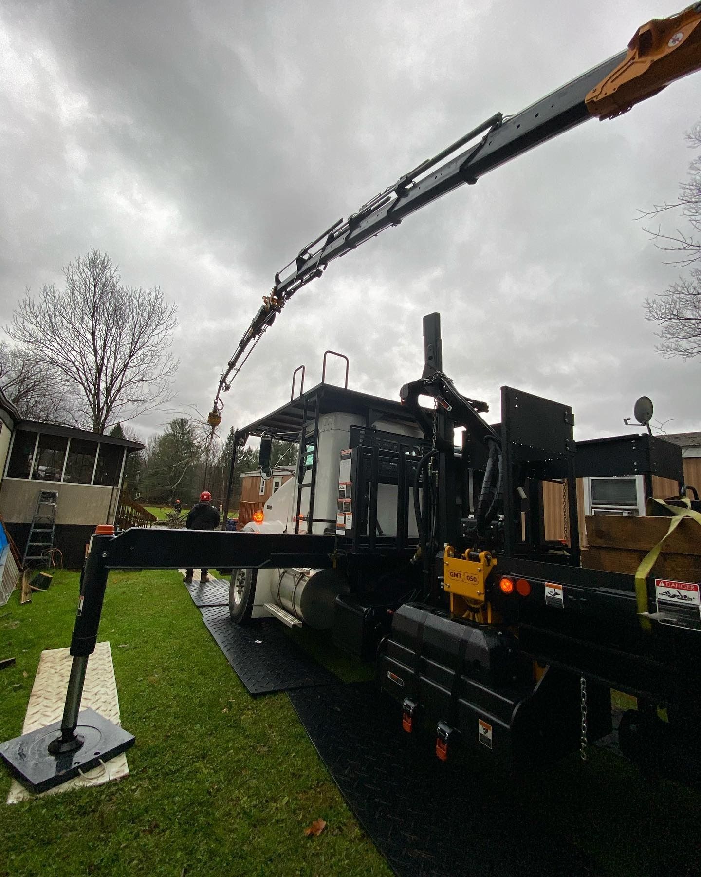 A large crane truck with an extended boom arm is parked on protective mats on a grassy lawn near a house.