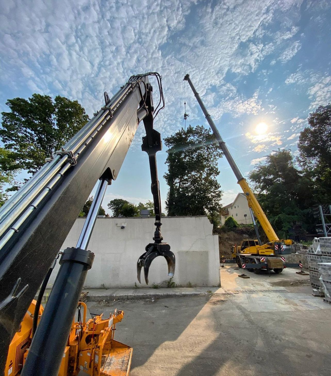 A yellow crane and a machine with a grapple attachment work at a construction site against a blue, cloudy sky.