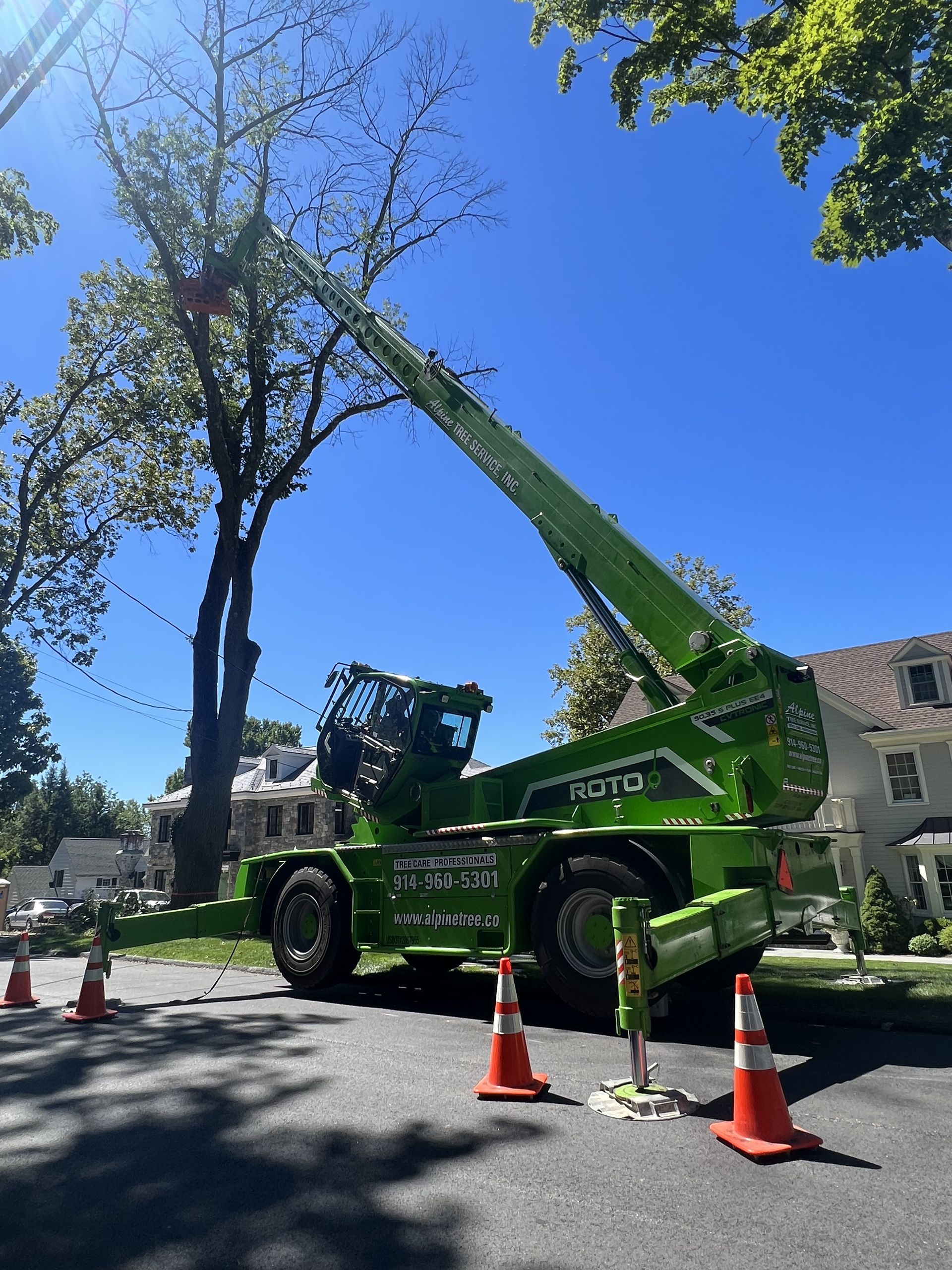A bright green crane truck with an extended boom arm is parked on a suburban street, parked near a large tree.