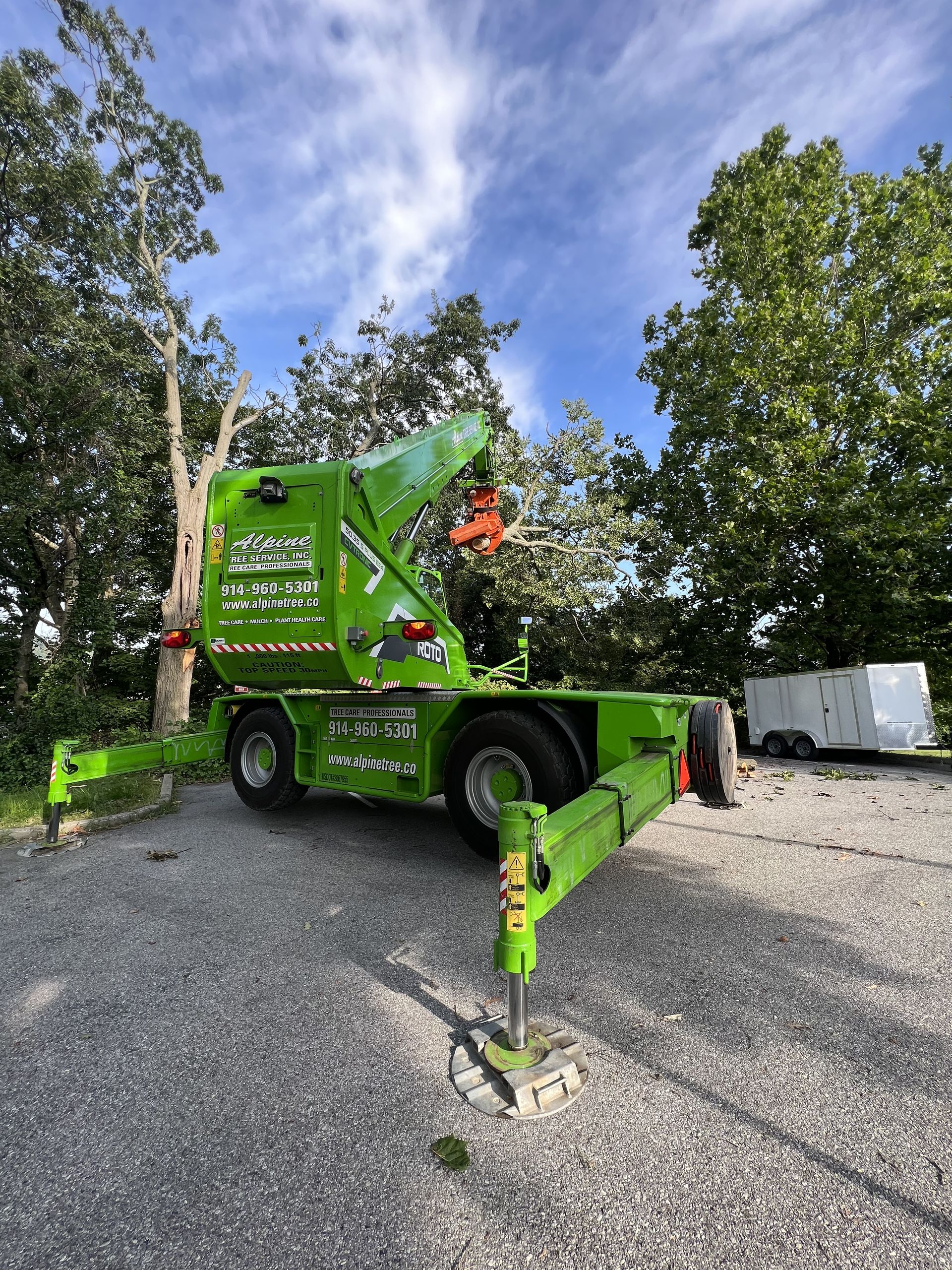 A bright green mobile crane with extended outriggers sits on a gravel lot in front of trees and a white trailer.