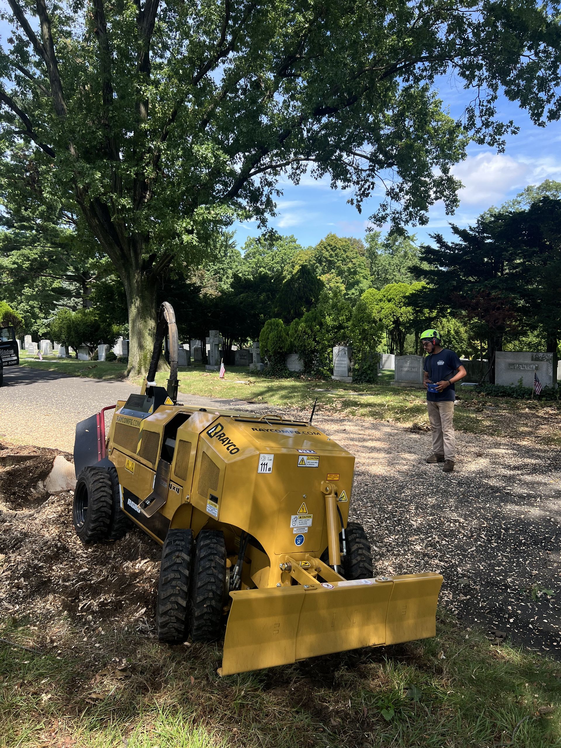 A person stands near a yellow ride-on trencher parked on gravel in a cemetery under large trees.