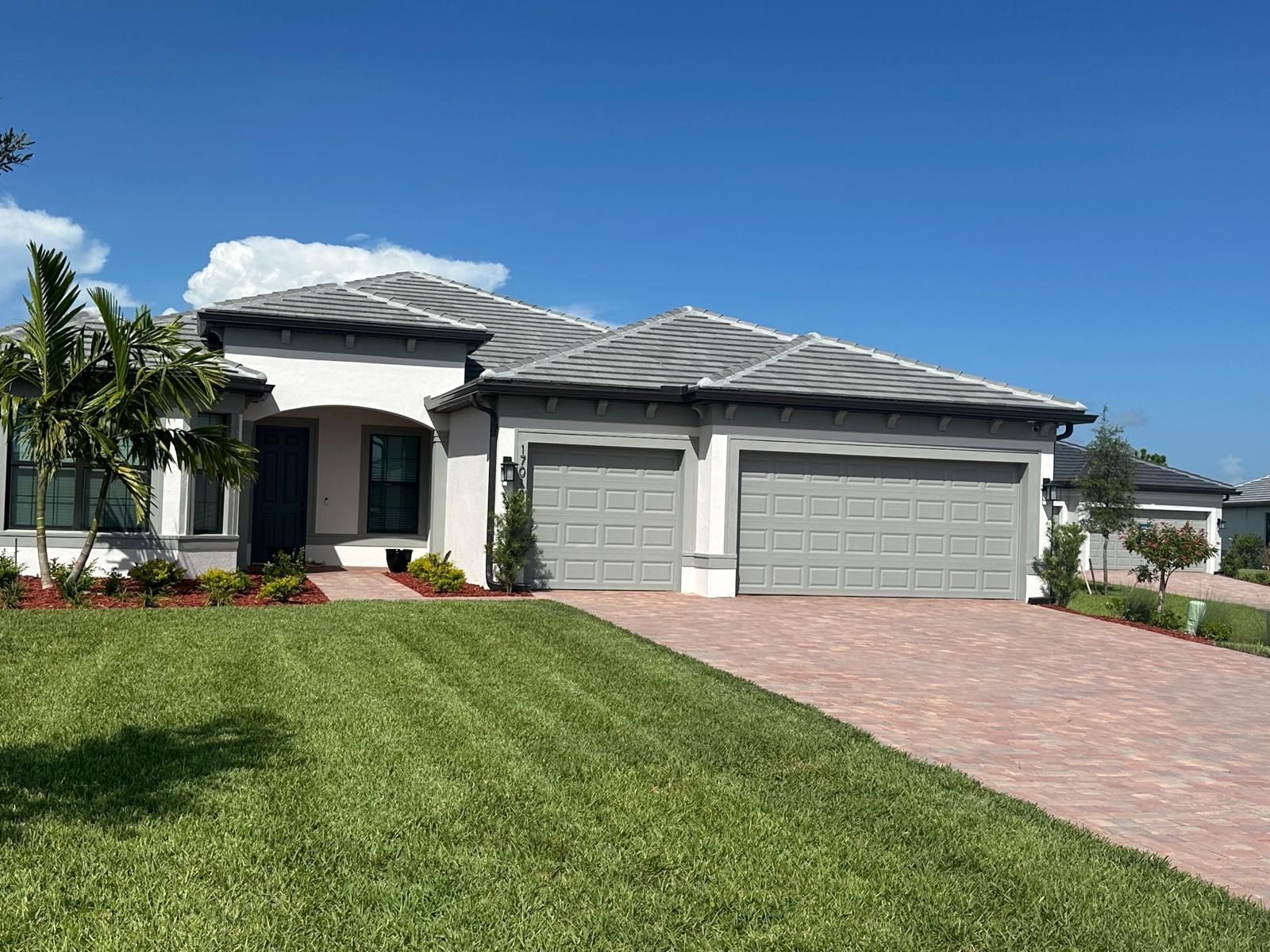 A light-colored house with a gray roof and two-car garage. Lush green lawn and a red brick driveway under a blue sky.