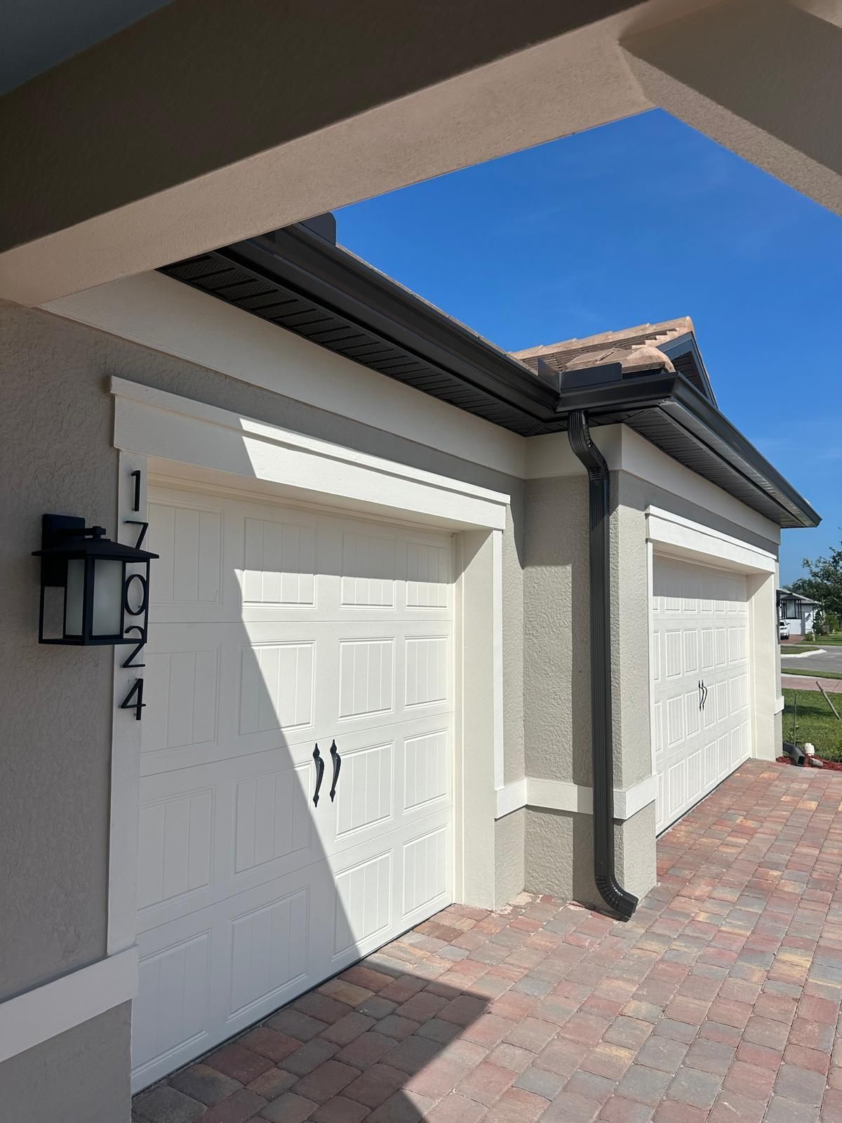 Exterior of a light-colored house with white garage doors, black gutters, and a street number. Clear blue sky.