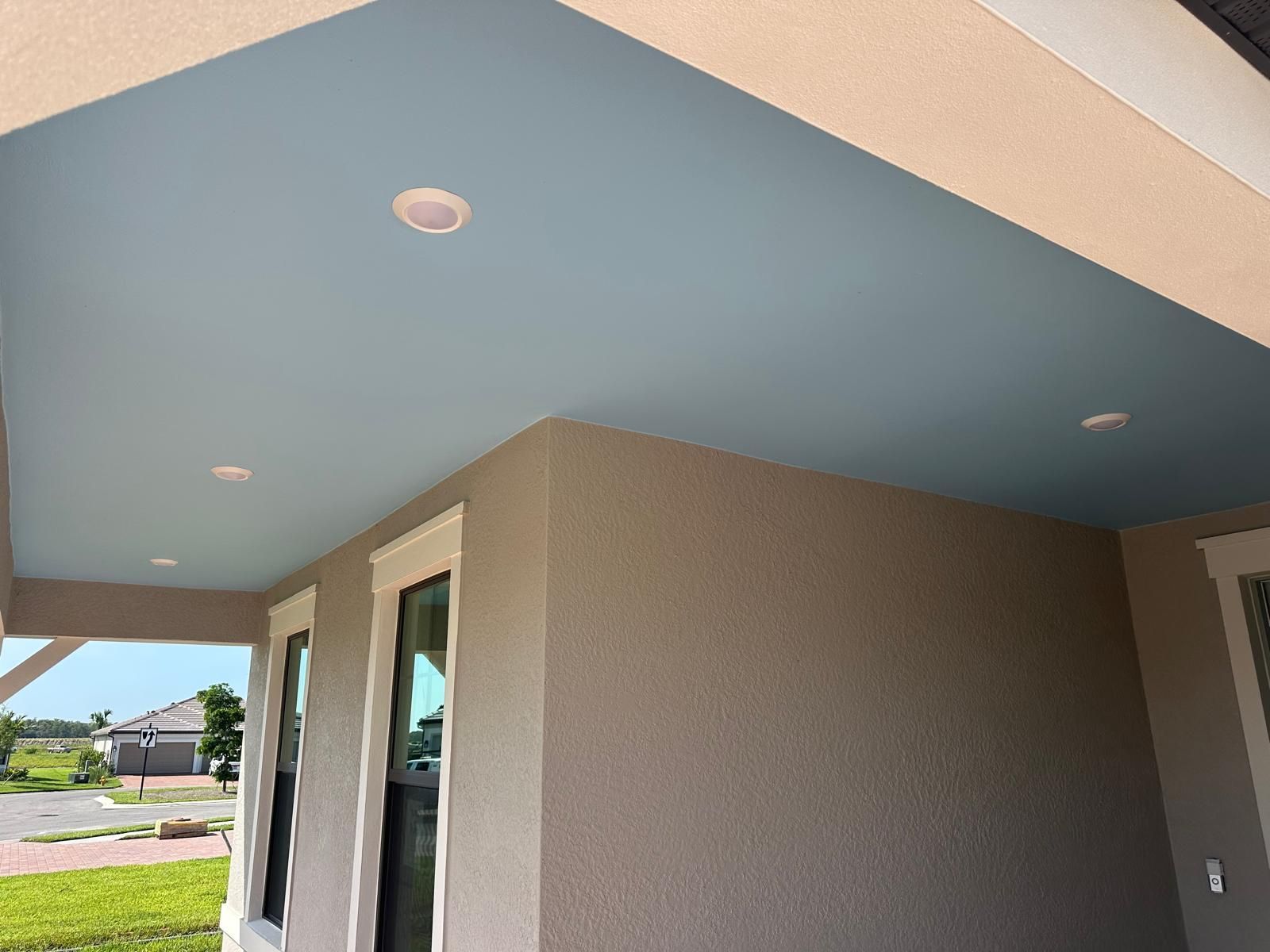 A porch ceiling painted light blue, with recessed lighting, and walls of a taupe color. Windows and a glimpse of a street are visible.