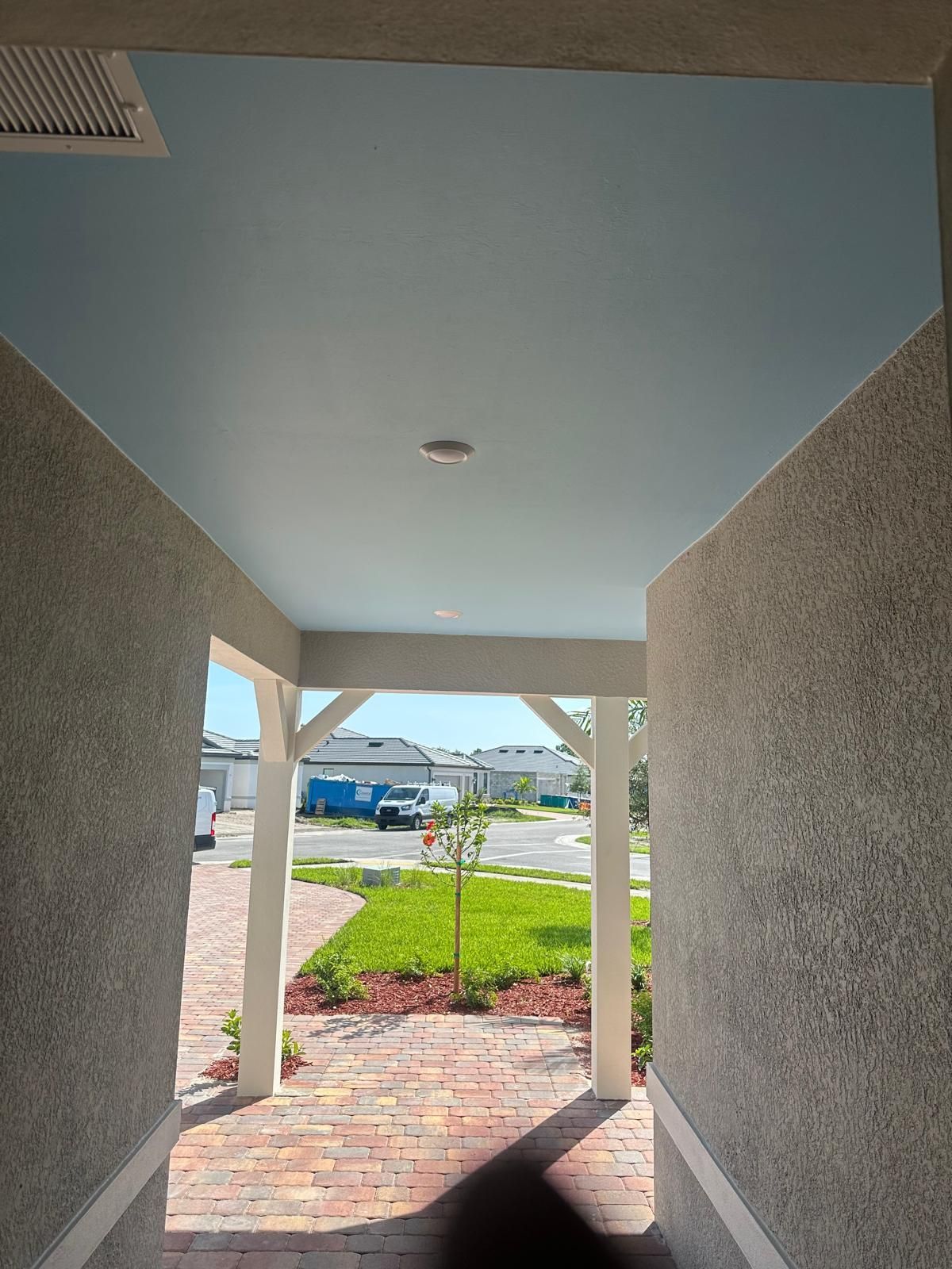 Looking out from under a covered porch with a light blue ceiling. Brick pathway leads to a yard with grass and trees.