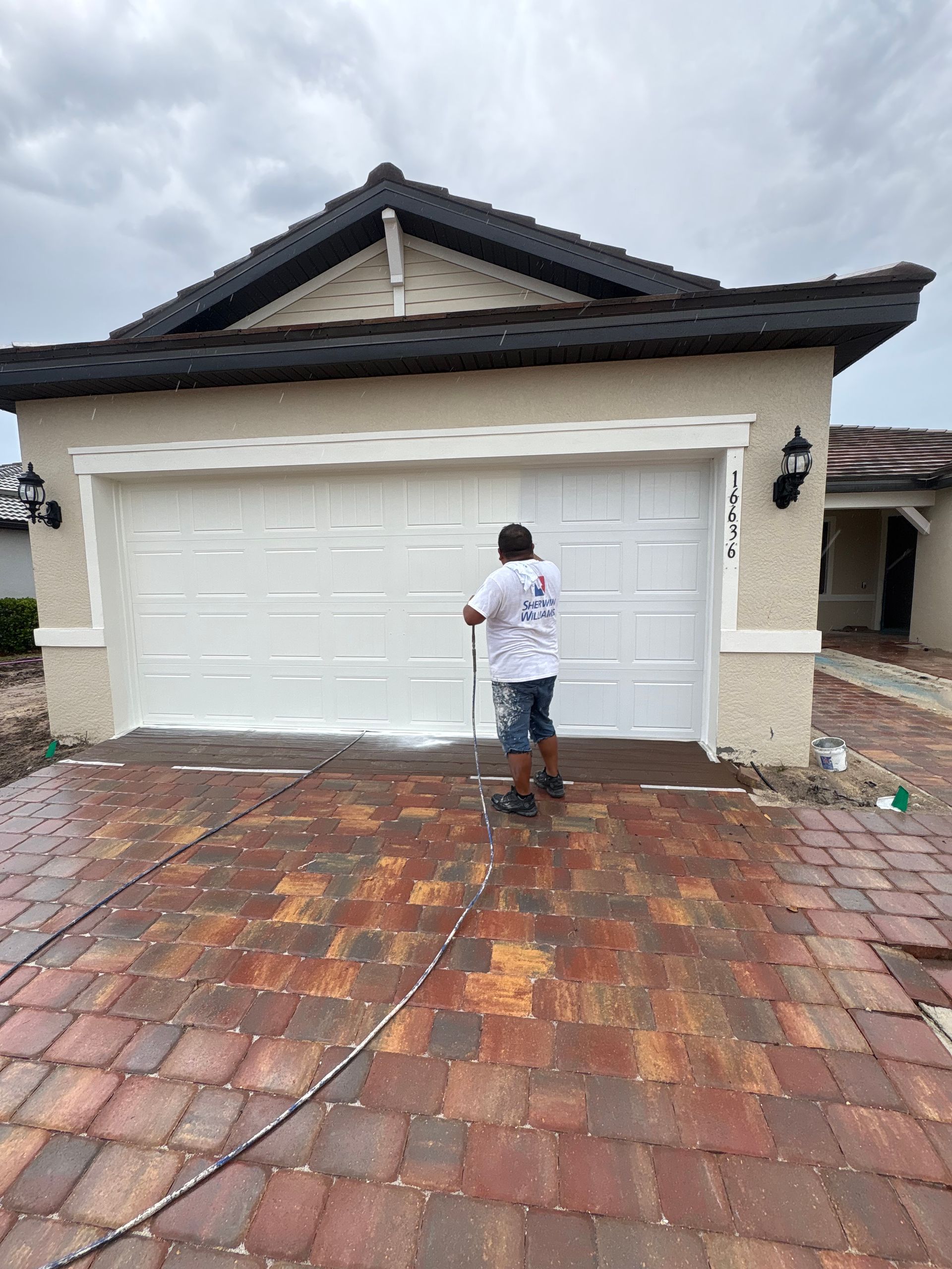 A person is pressure washing a white garage door of a house with a brick driveway on a cloudy day.