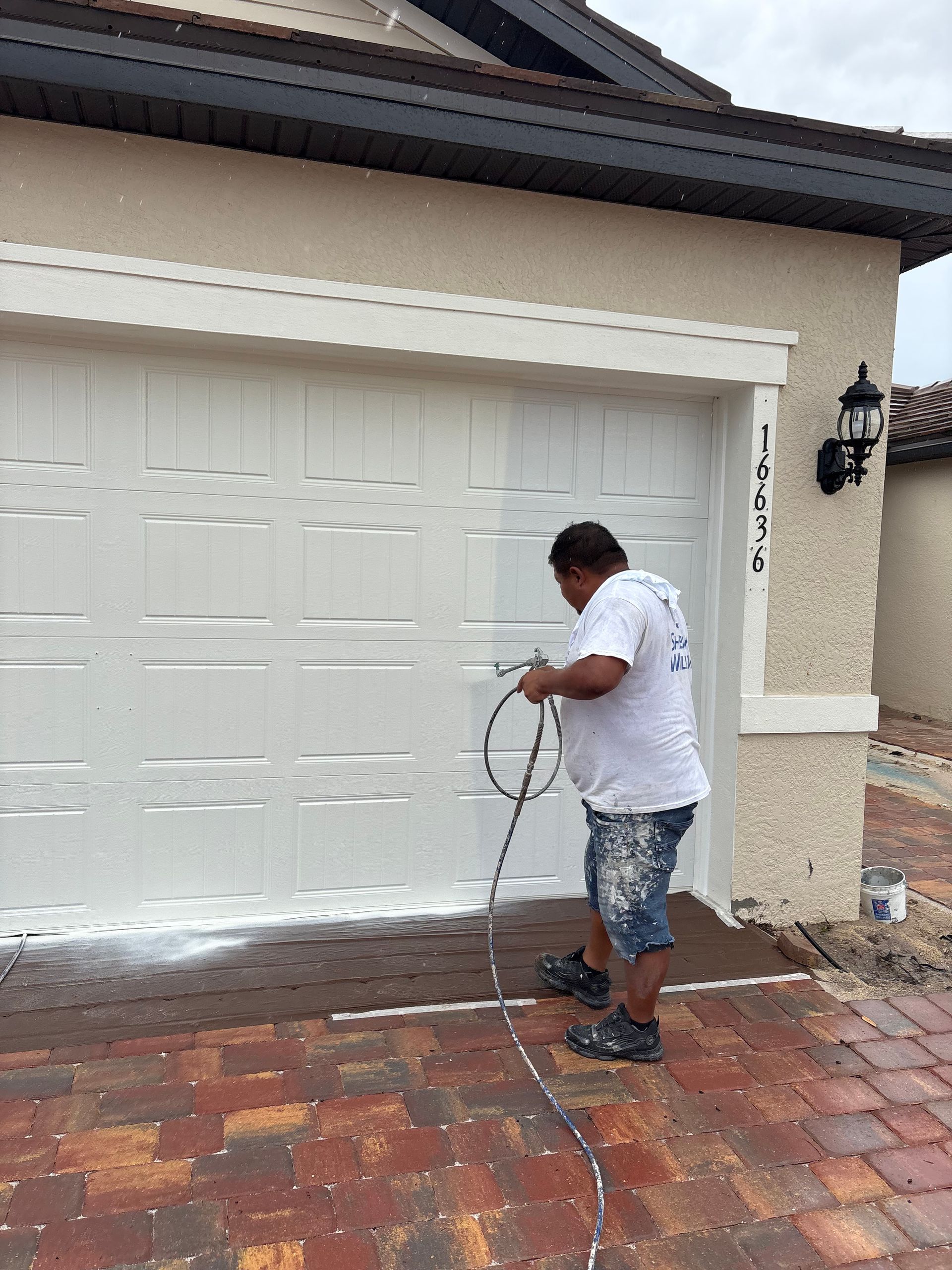 A person is painting a white garage door with a paint sprayer, standing on a brick driveway.