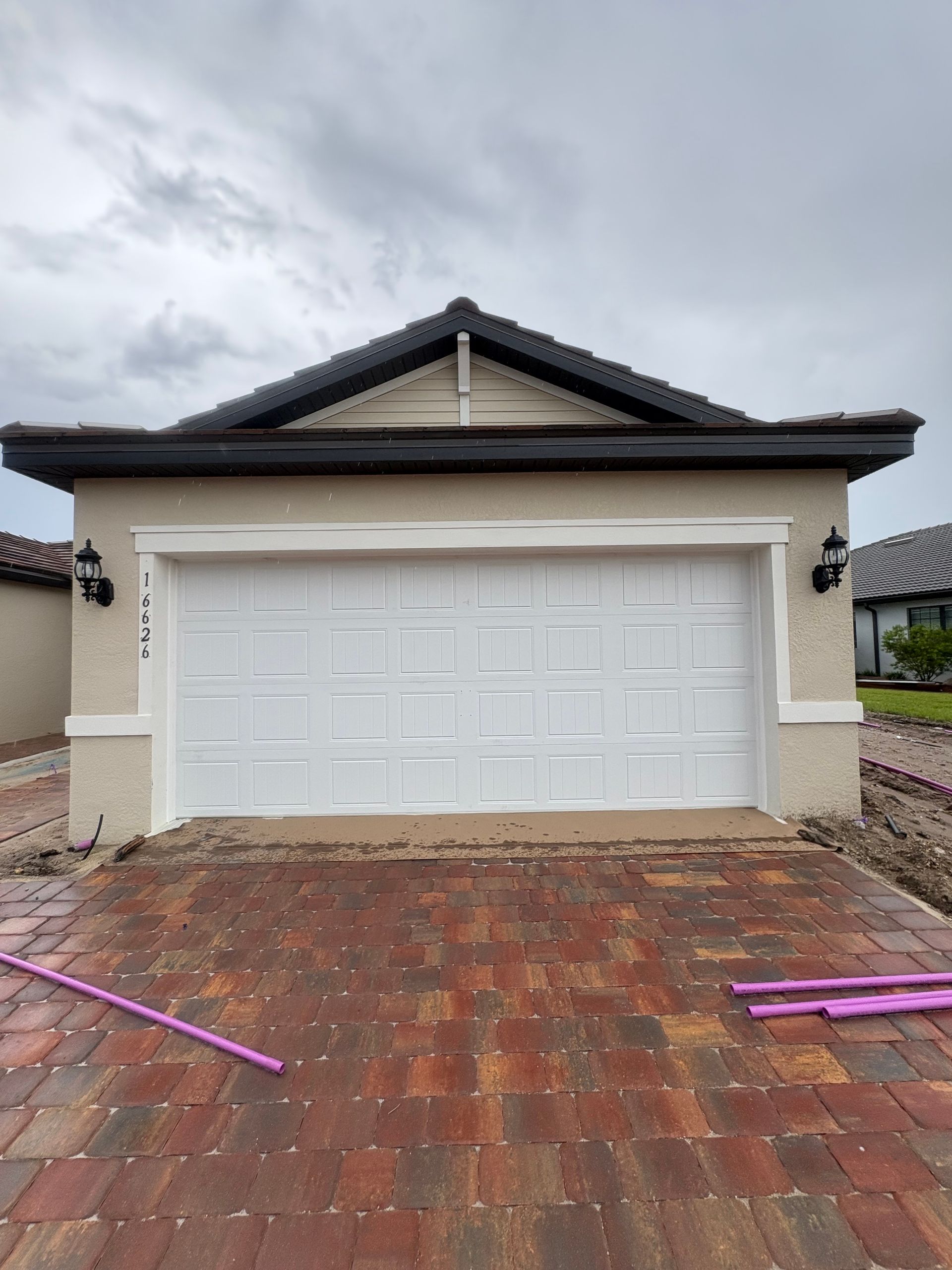 White garage door on a beige house with black trim, set on a brick driveway, under a cloudy sky.