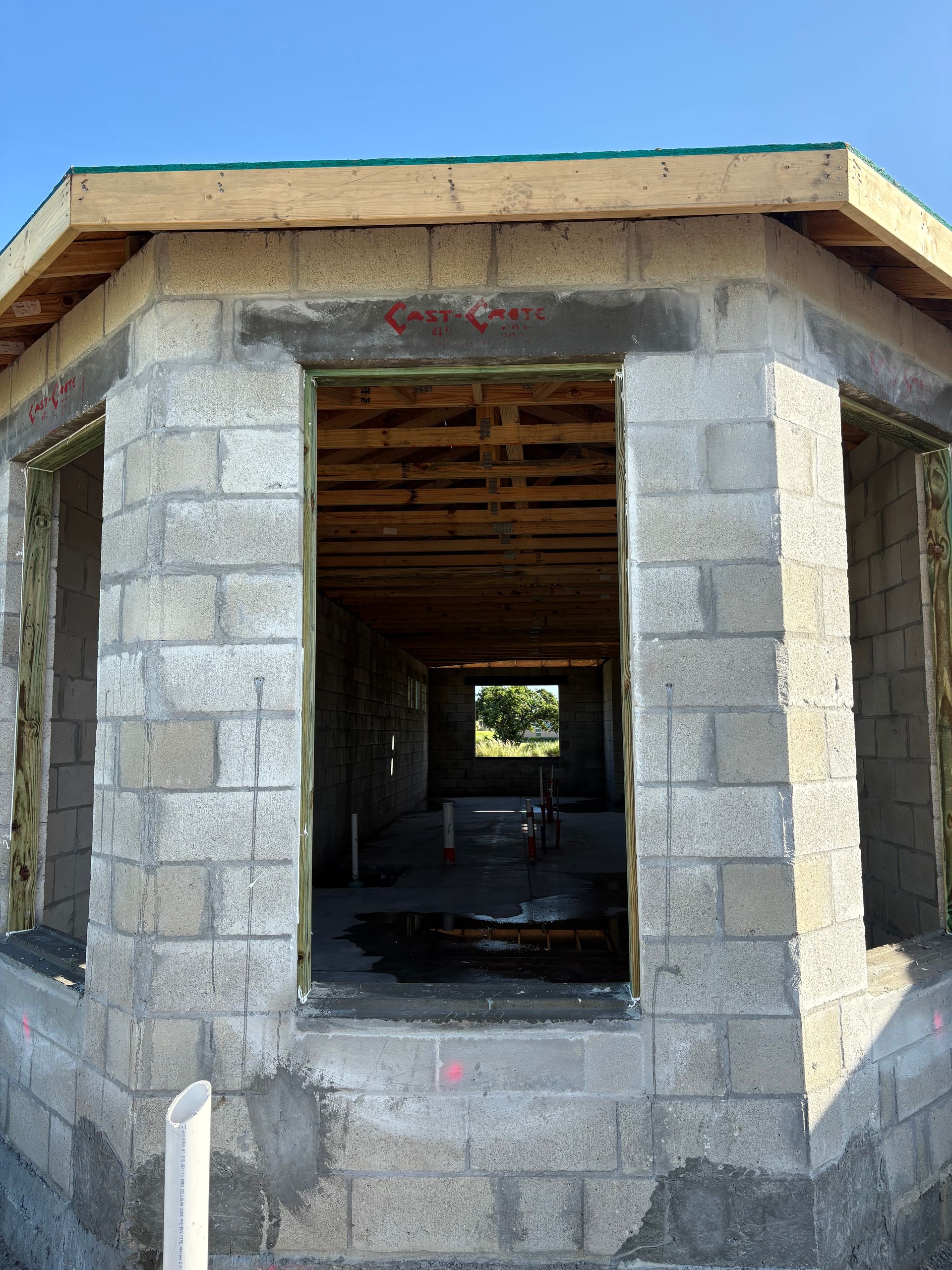Exterior of a building with concrete block walls, window openings, and exposed wooden beams at the roofline, against a blue sky.