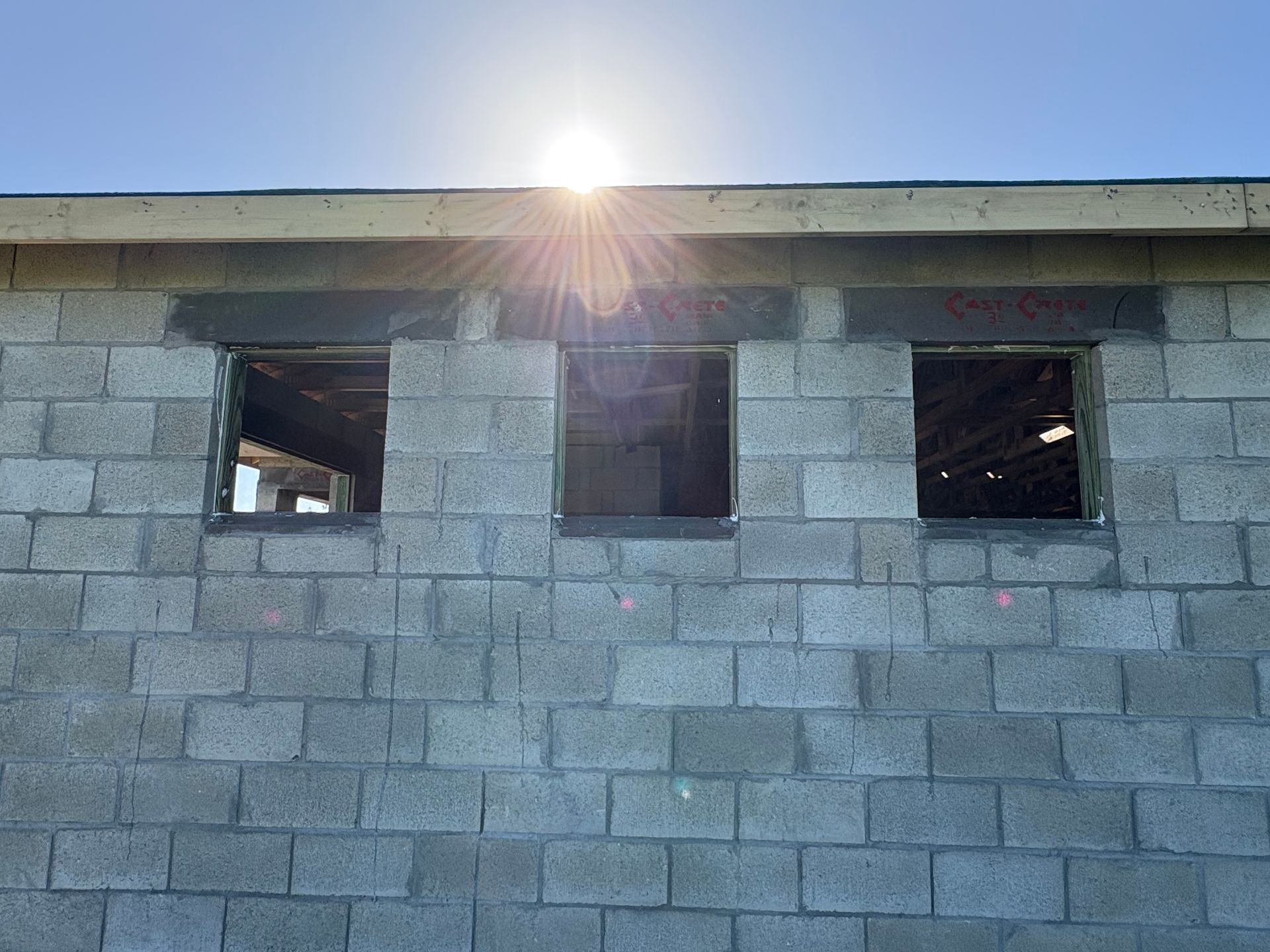 Exterior of a cinder block building under construction, three window openings beneath the sun.