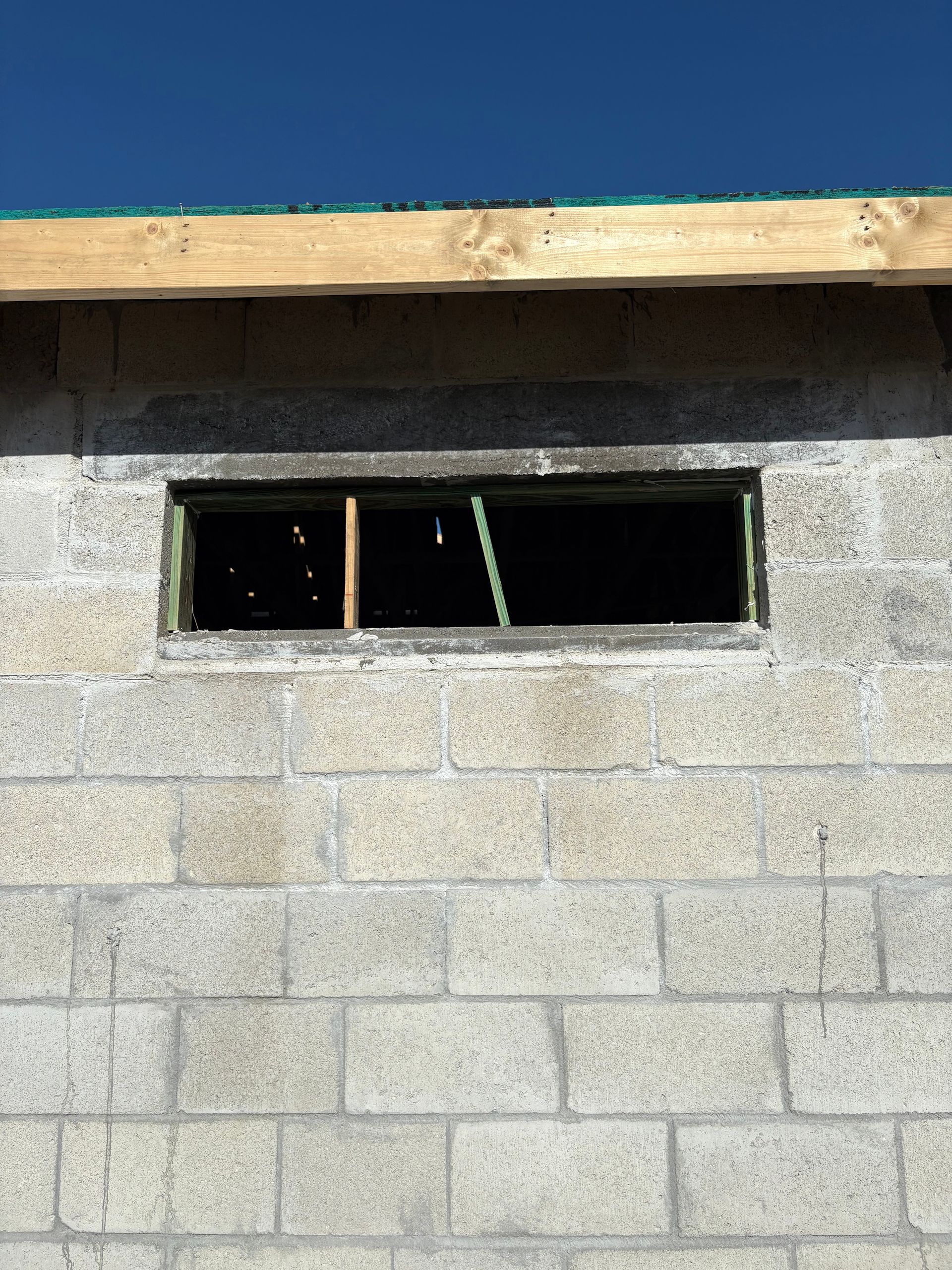 Window opening in a gray cinder block wall, under a wooden beam and blue sky.