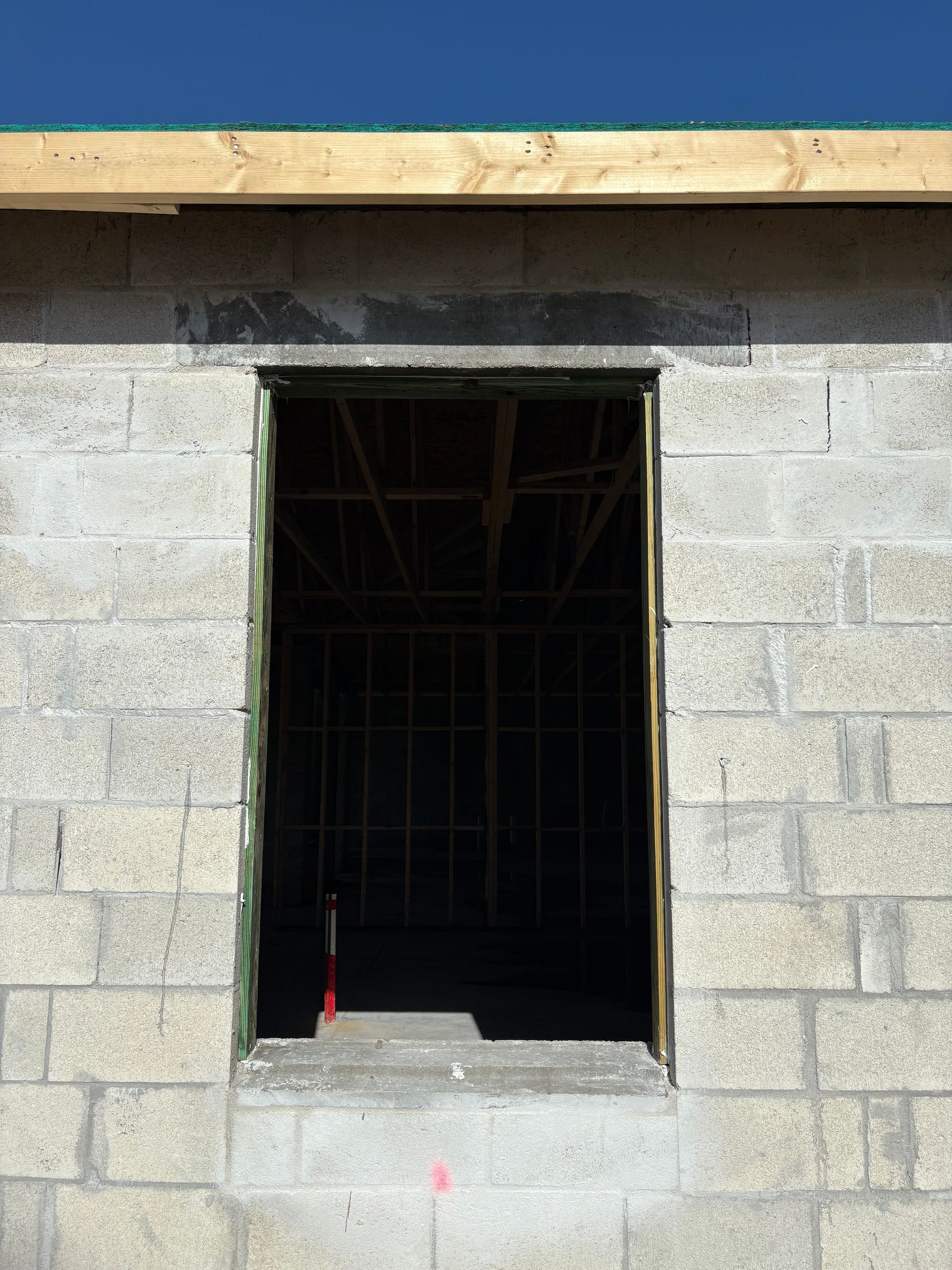 Exterior view of a cinder block building under construction. A rectangular opening for a doorway or window is visible against a bright blue sky.