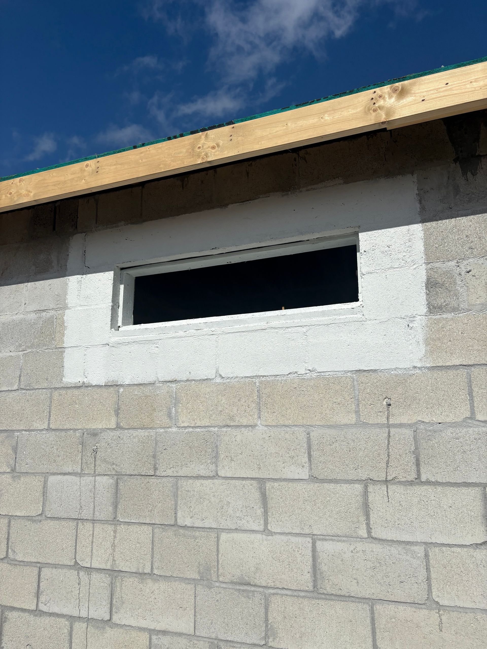 Exterior view of a concrete block wall with a rectangular window. The window has white trim and sits beneath a wooden beam.
