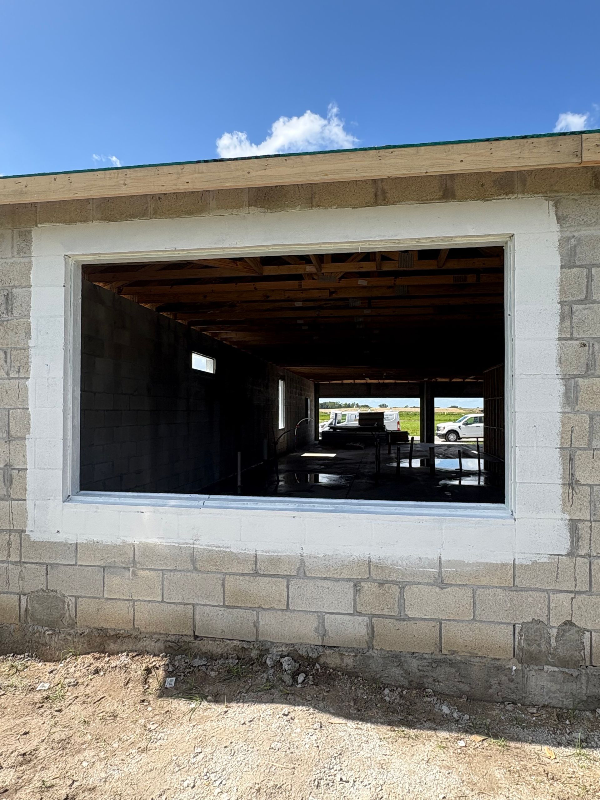 Exterior of a cinder block building with a large window frame painted white. The open window reveals the interior and a view to the outside.
