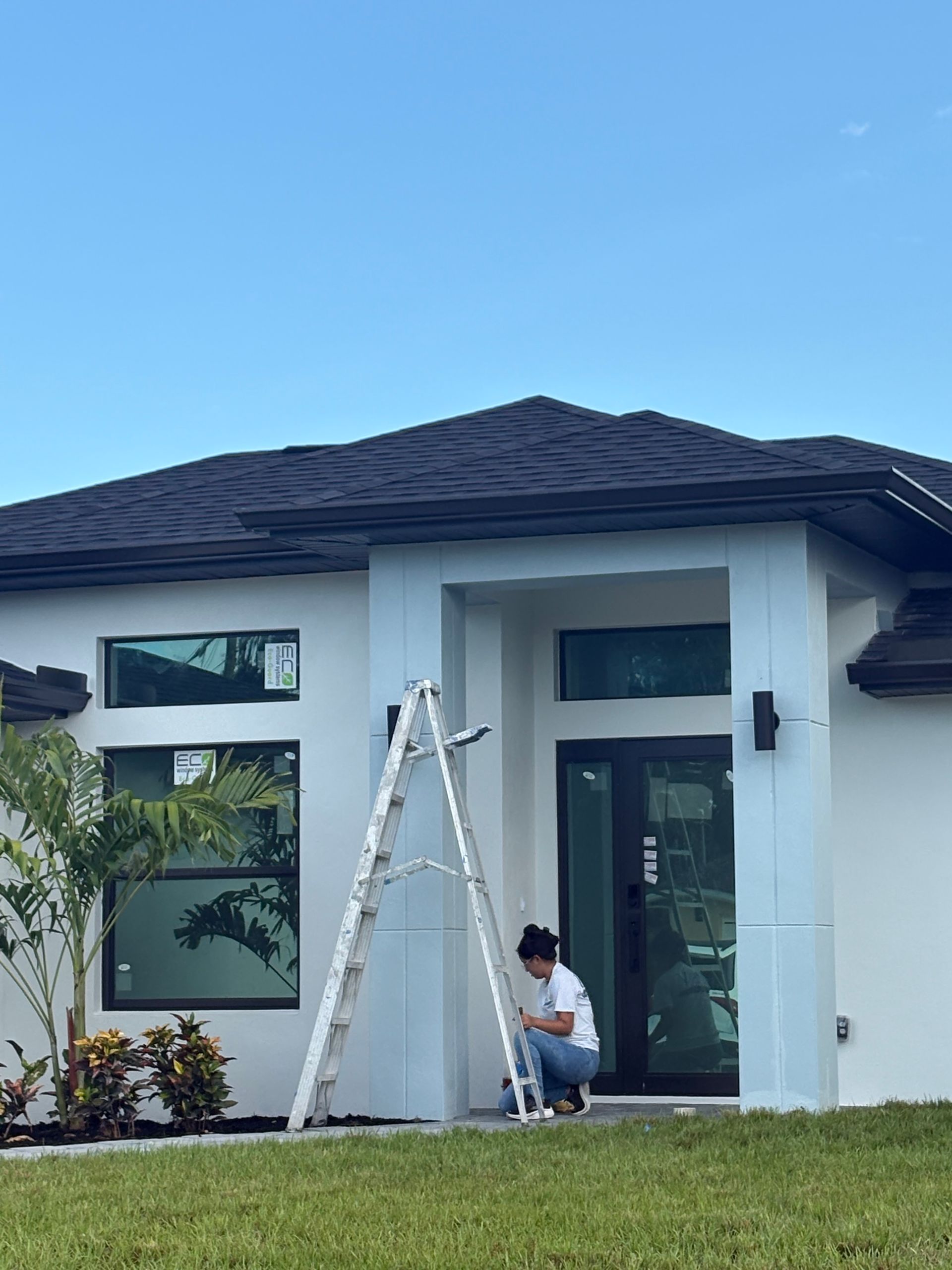 Person on a ladder painting the exterior trim of a white house. A sunny, blue sky is above.