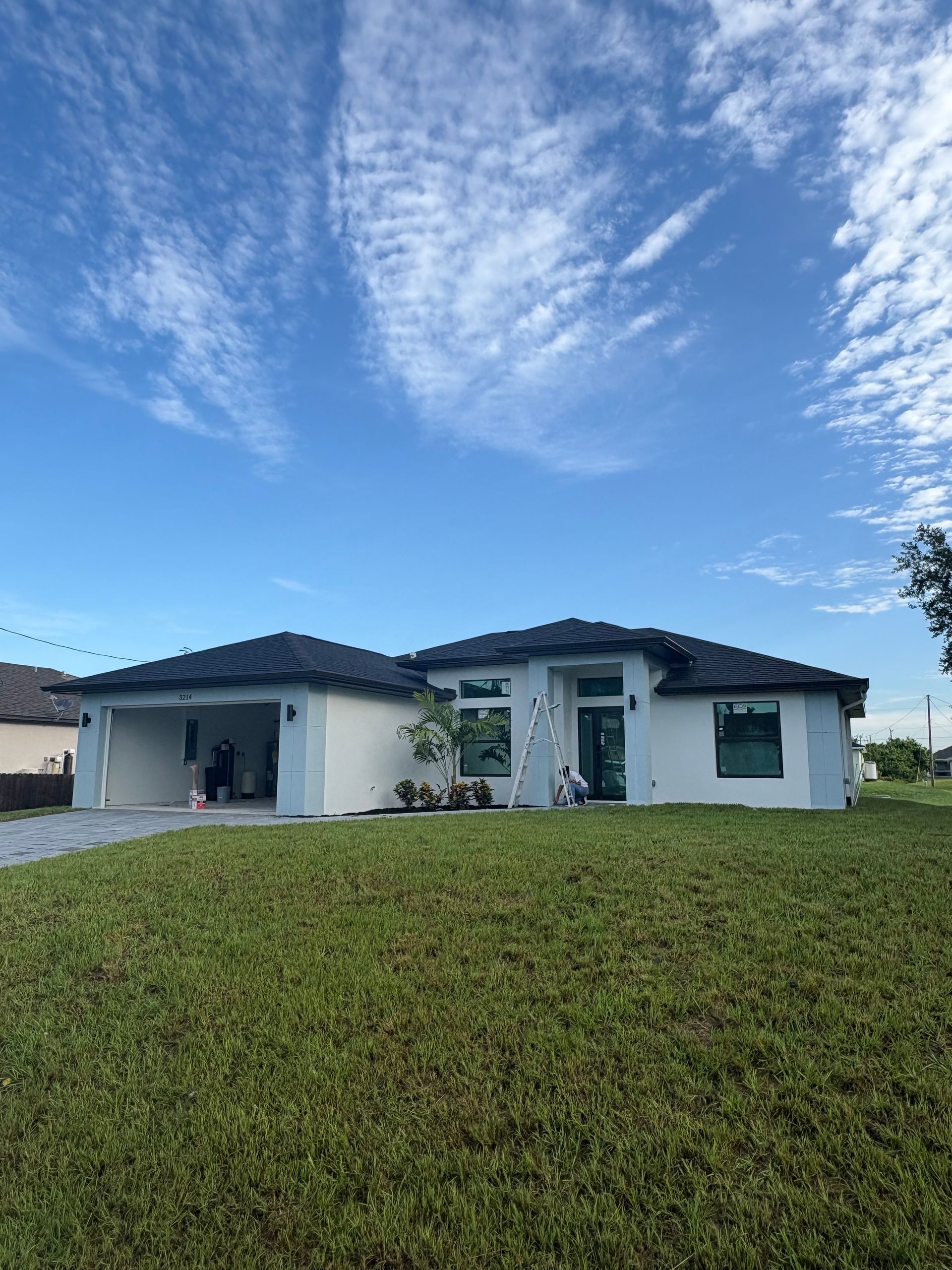 White house with dark roof and attached carport on a green lawn under a blue sky with streaky clouds.
