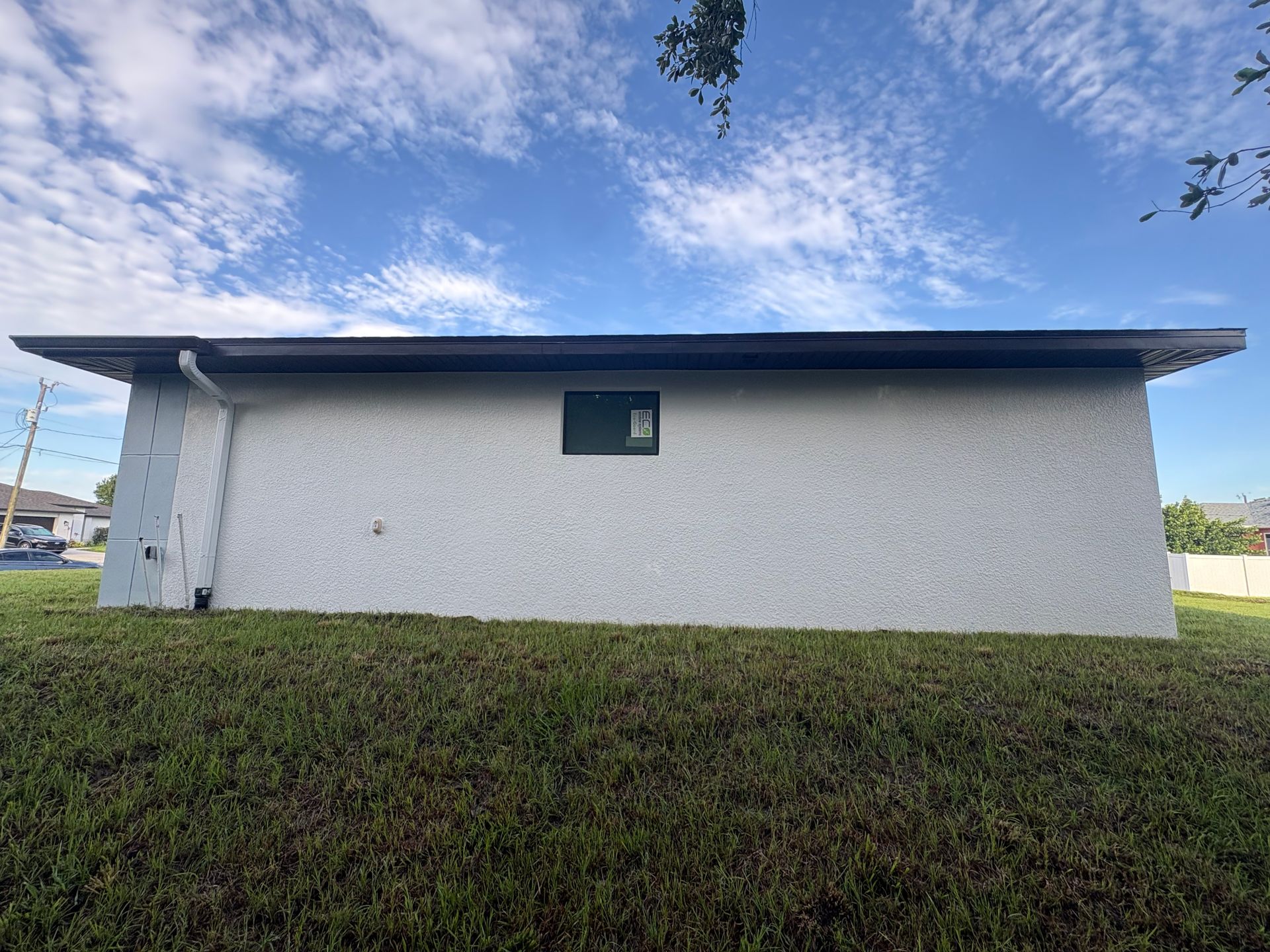 Side view of a stucco building with a dark roof, a small window, and a grassy lawn under a cloudy sky.