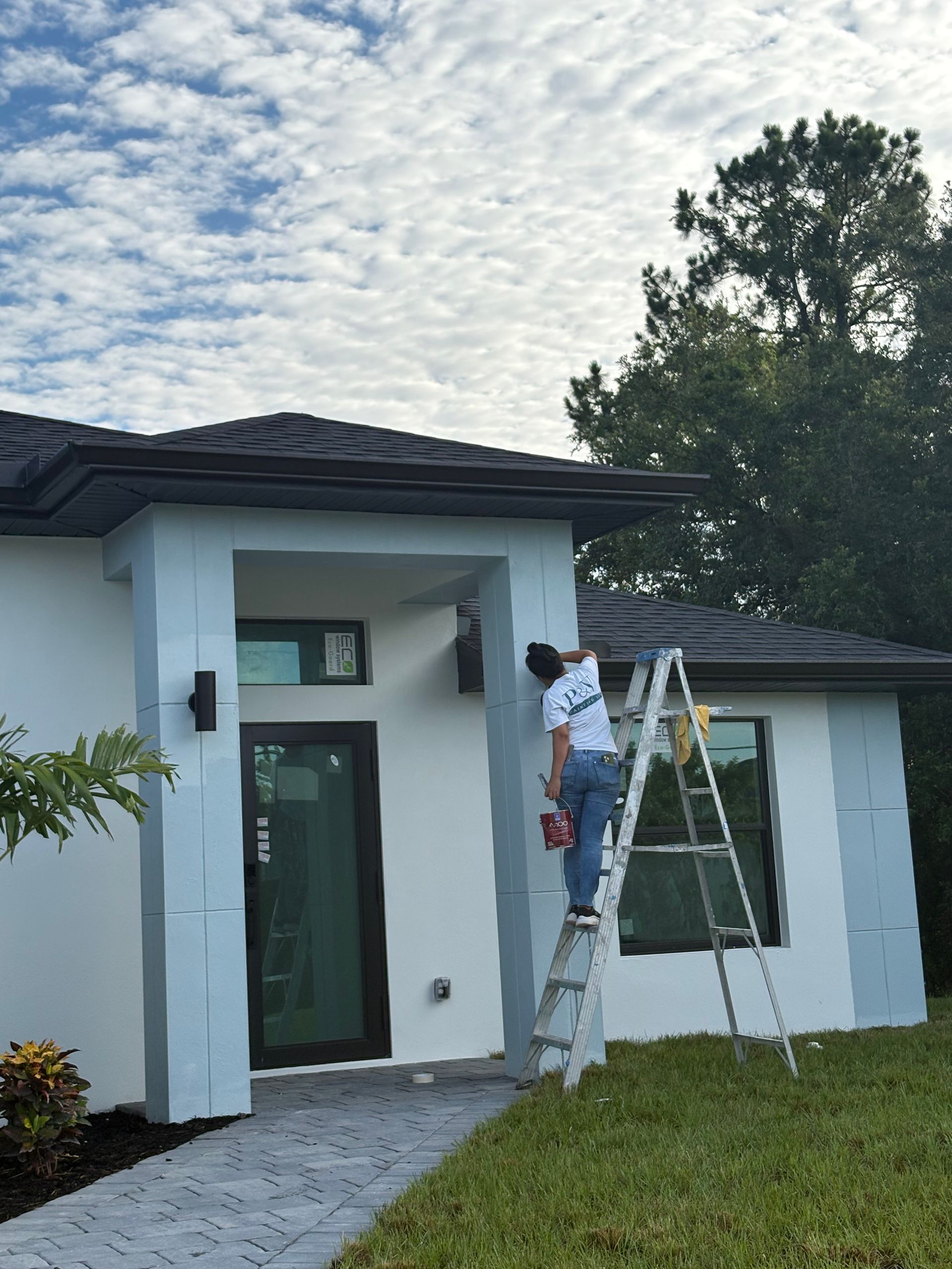 A person on a ladder paints the exterior of a modern, light-blue and white house under a cloudy sky.