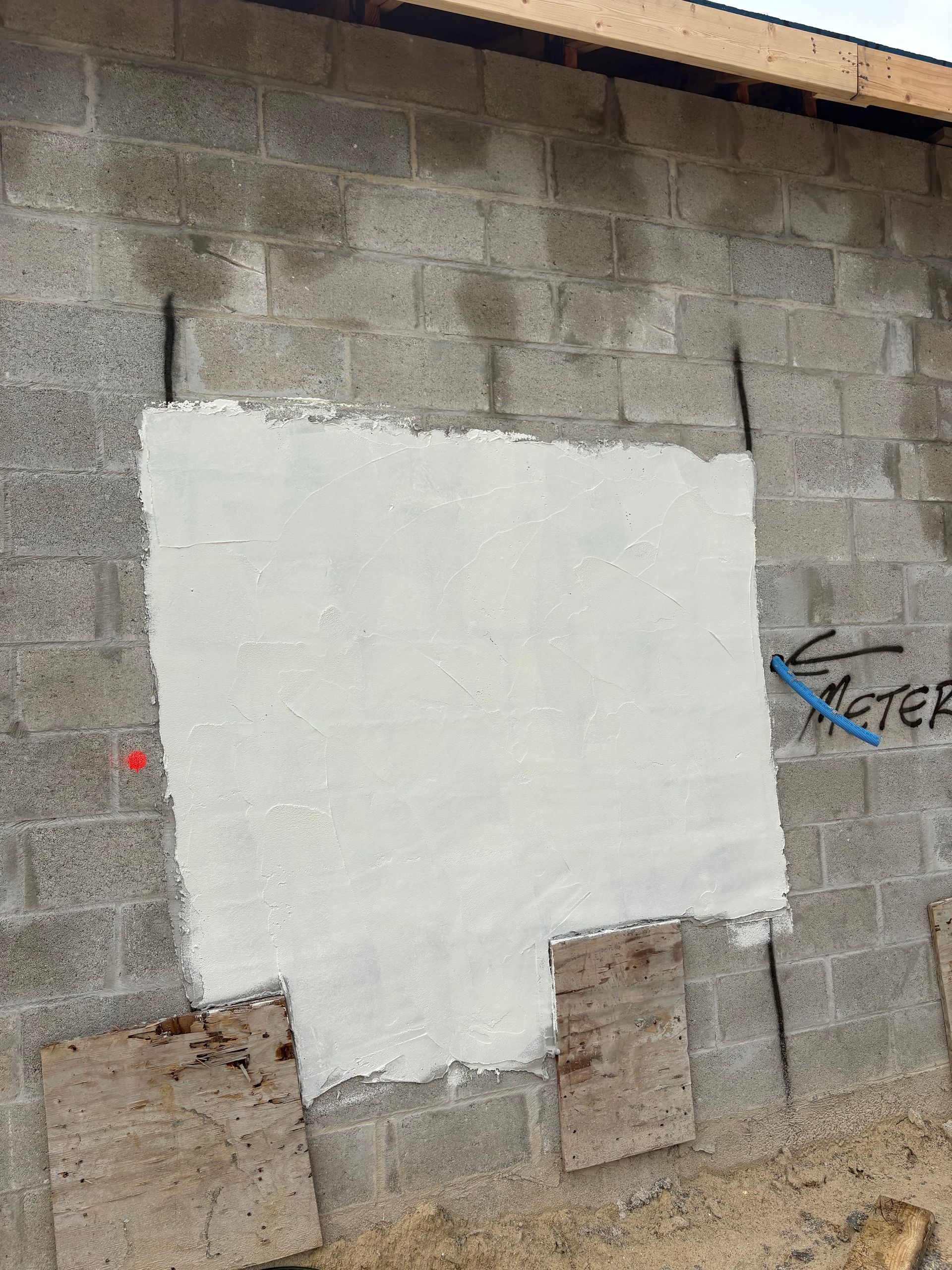 Gray cinder block wall with a patch of white cement, wood panels below. Blue marker indicates a water meter.