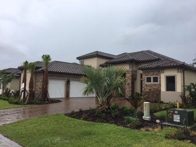 A two-story beige house with a brown tile roof, white garage doors, and stone accents sits in a grassy yard on an overcast day.