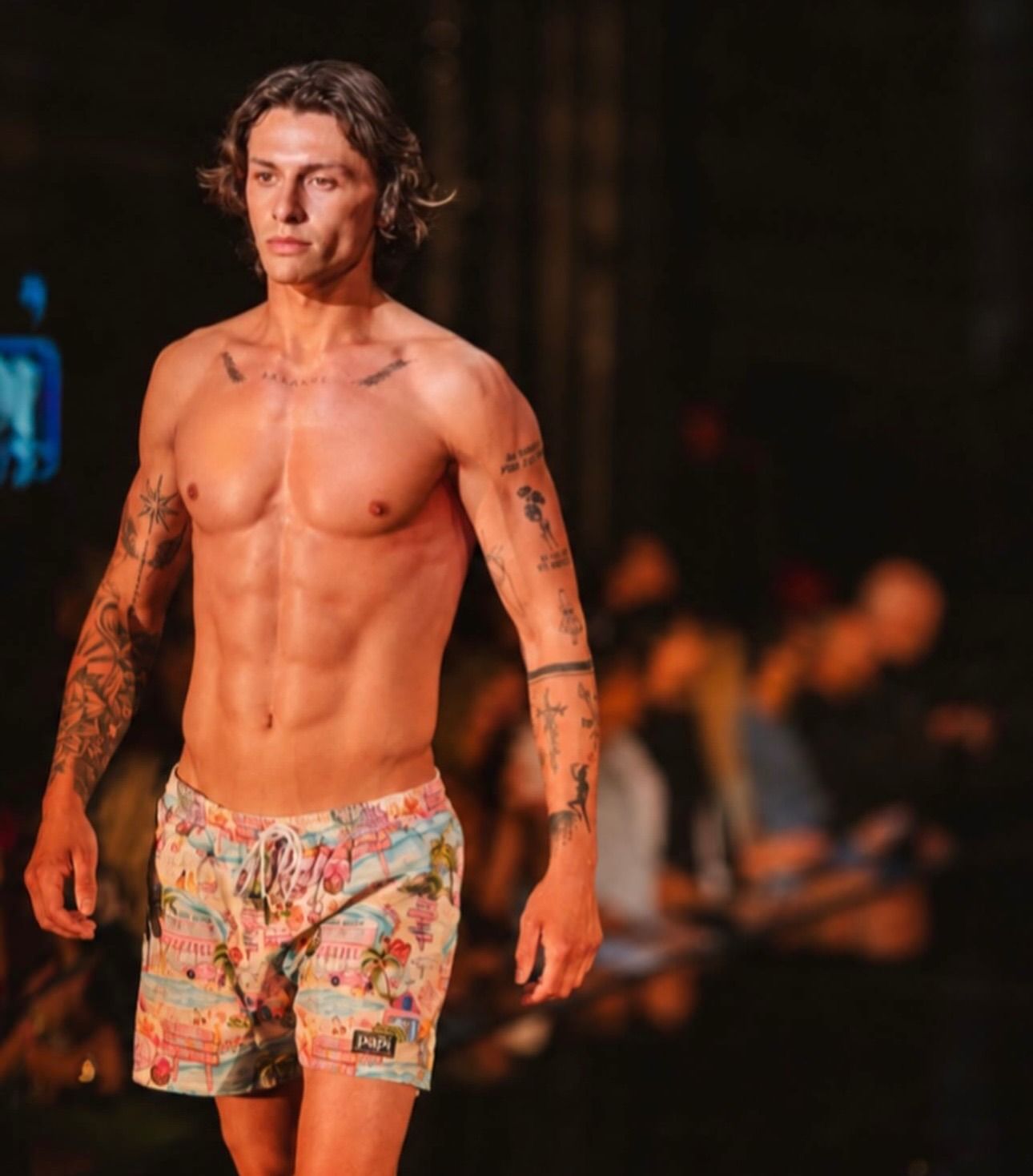 Shirtless model walking a runway in floral shorts, with tattooed arms and warm stage lighting