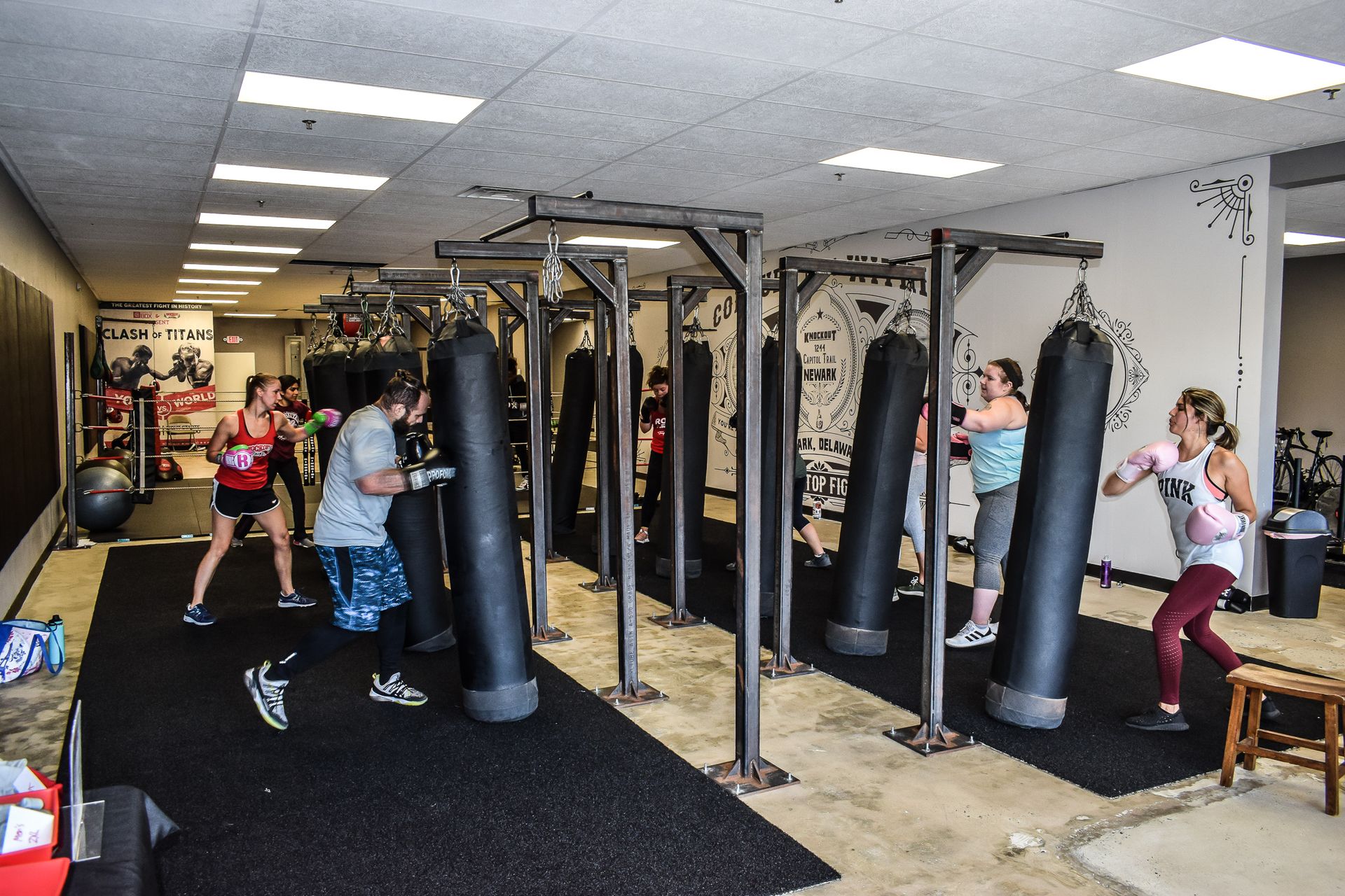 People boxing at a fitness studio. They punch heavy bags. Black mats, equipment, and light-colored walls.